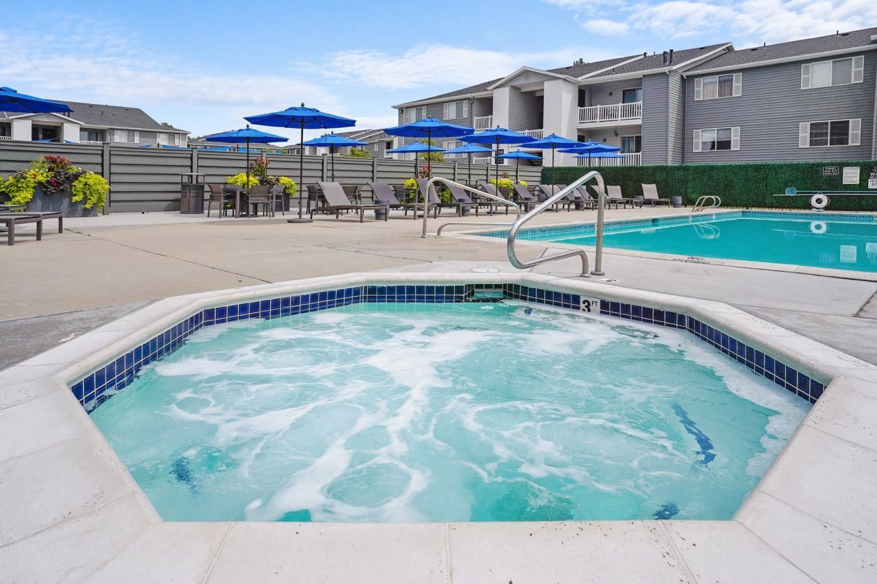 Outdoor community pool with a hot tub in the foreground, blue umbrellas, and lounge chairs.