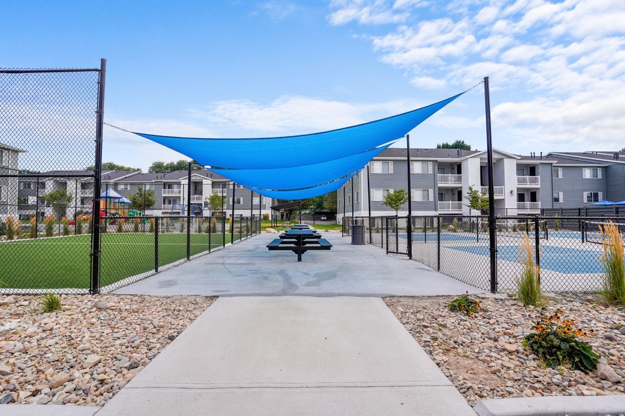 Outdoor community amenity area with a blue shade sail over picnic tables and a fenced recreation court.  Explore spacious Millcreek townhomes for rent by visiting Turnberry Apartments for a community tour.
