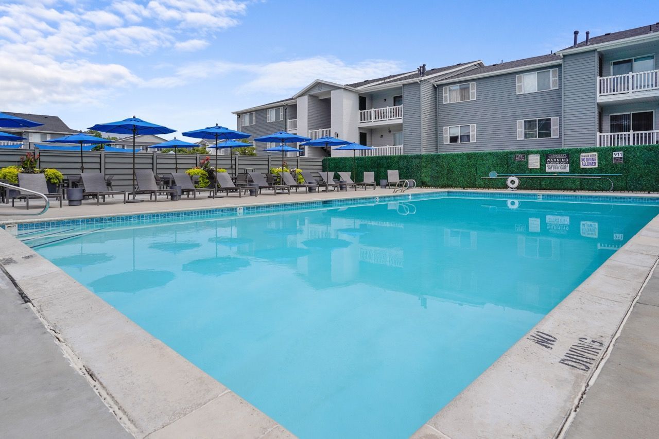 Outdoor pool at an apartment community with blue umbrellas and lounge chairs along a concrete deck.