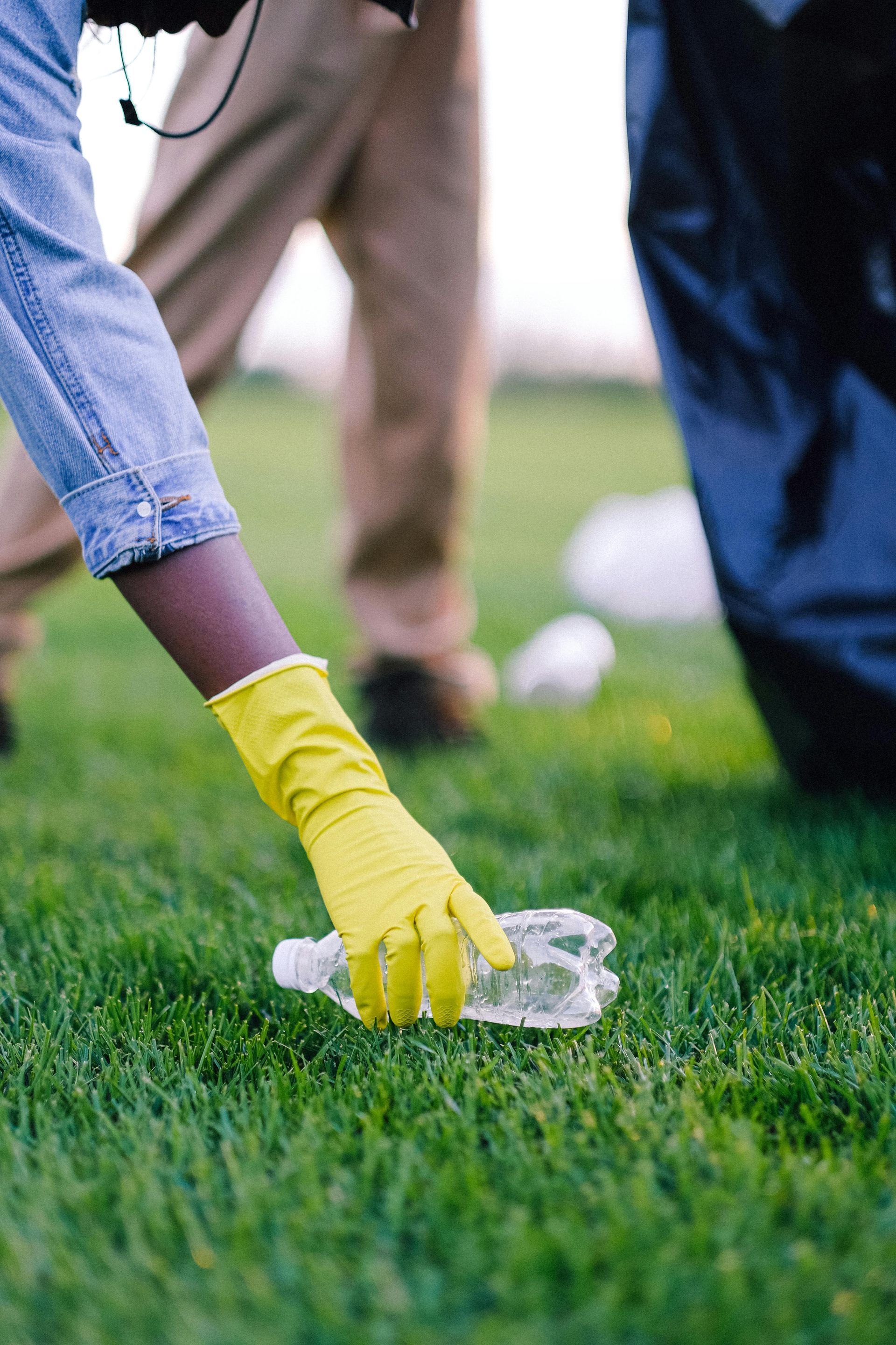 A person wearing yellow gloves is picking up a plastic bottle on the grass.