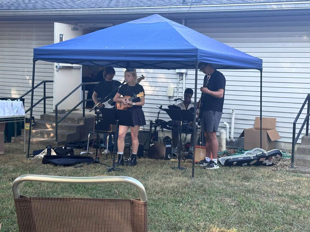 Band playing under a blue tent outdoors. Instruments include guitar, ukulele, and keyboard.