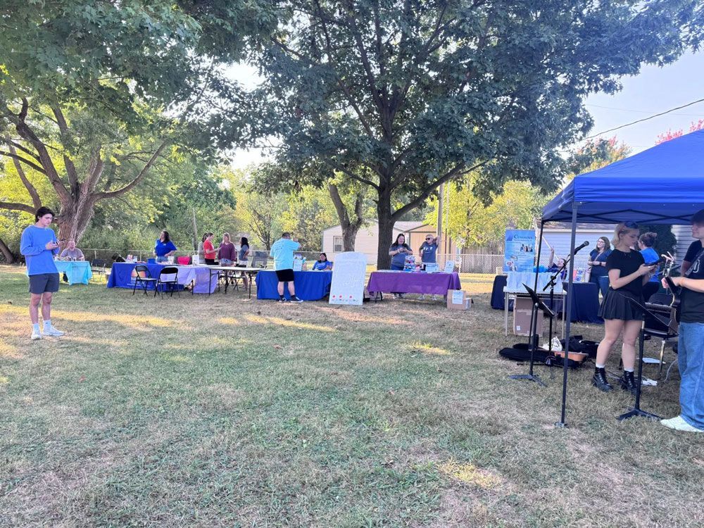 Outdoor event with tables, a band performing, and people in a park setting under a tree, with blue and purple canopies.