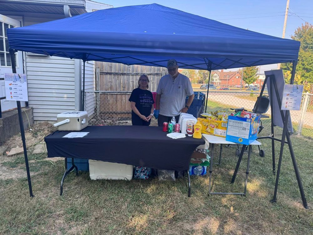 People at a table under a blue canopy; giving snacks and drinks outside, on a sunny day.