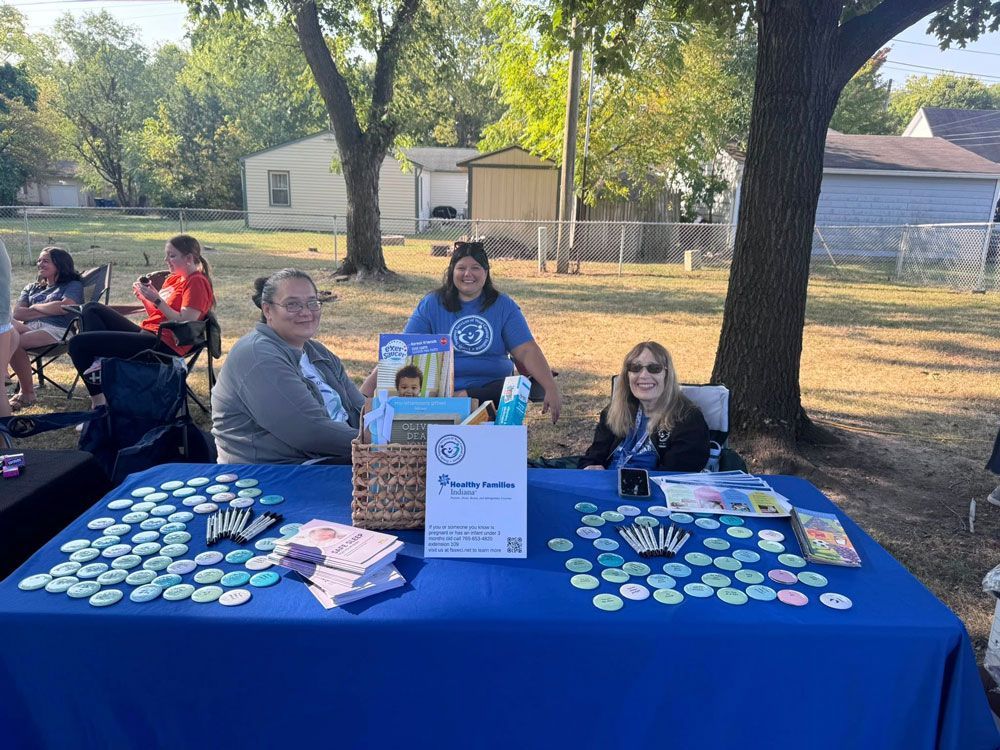 Three people at a table, with items outdoors. Blue tablecloth with products and a sign.