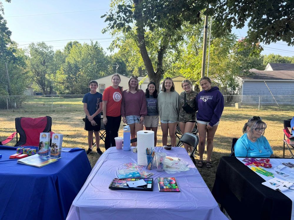 Group of people with tables displaying items. Trees and a house in the background.