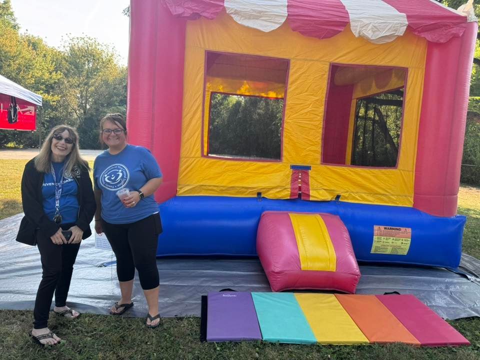 Two women stand by a colorful bounce house on grass, outdoor setting.