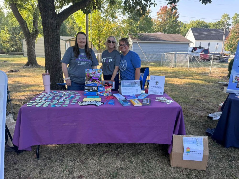 Three women stand behind a purple table at an outdoor event, with promotional items and materials on display.