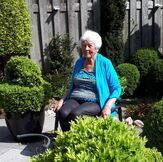 Een oudere vrouw met wit haar lacht in een tuin, omringd door groene struiken, gekleed in een blauwe trui.