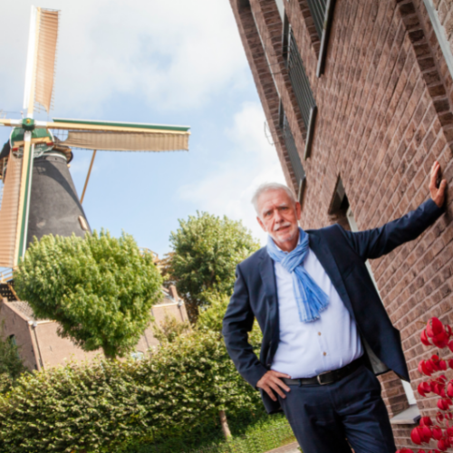 Man in pak, sjaal, leunend tegen een bakstenen gebouw bij een windmolen. Blauwe lucht, groen gebladerte.