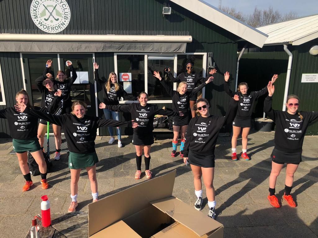 Een hockeyteam in zwarte shirts en rokken juicht voor het clubhuis.