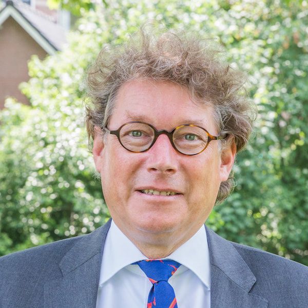 Een man met een bril lacht, gekleed in een pak en een stropdas met print, buiten in het groen.