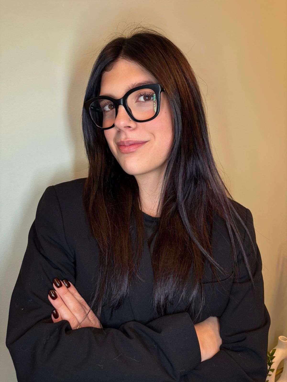 Donna con lunghi capelli scuri, occhiali e blazer, braccia incrociate, sorridente.