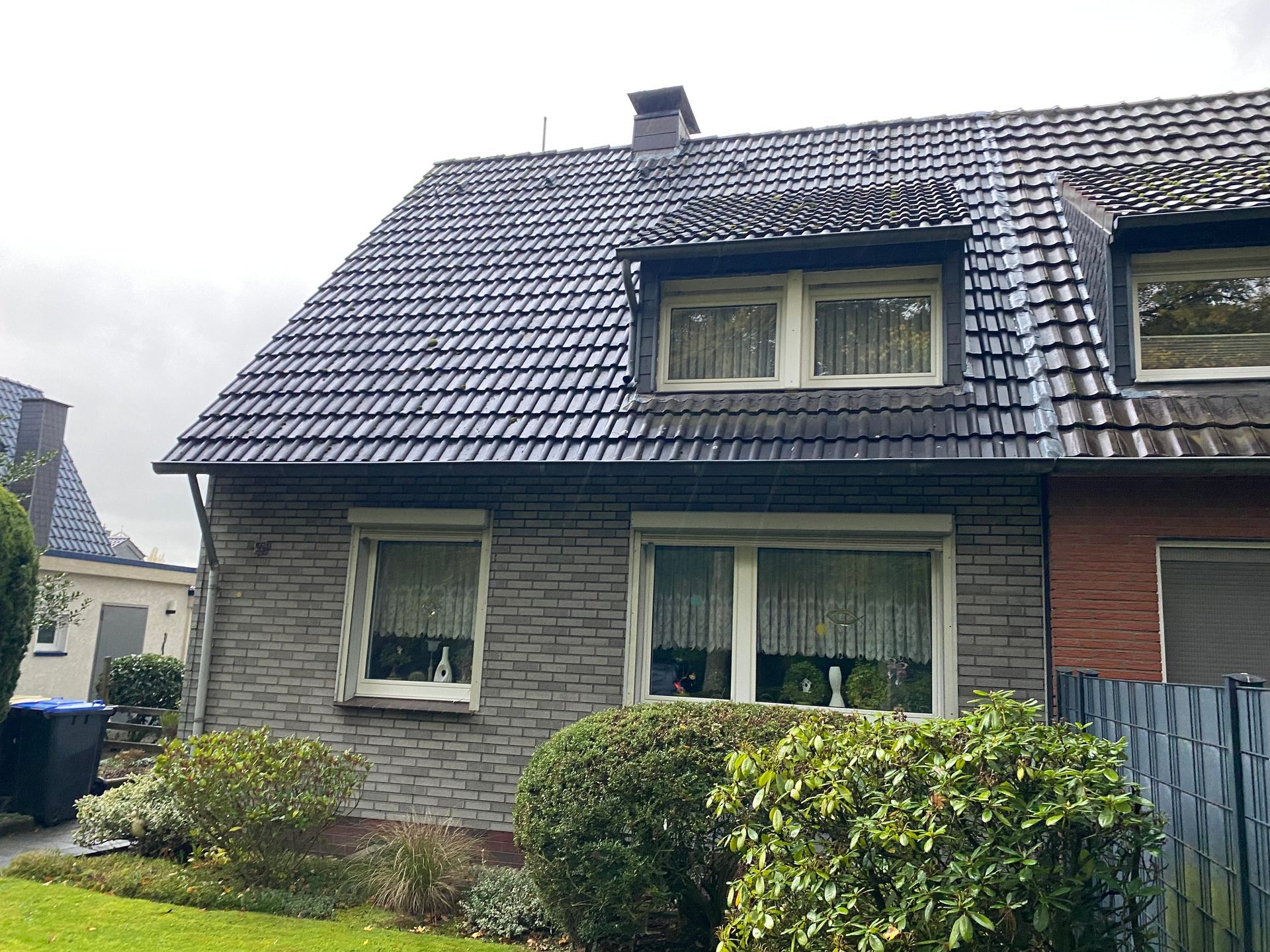 Graues Backsteinhaus mit dunklem Ziegeldach, Fenstern und grünen Büschen. Bewölkter Tag.