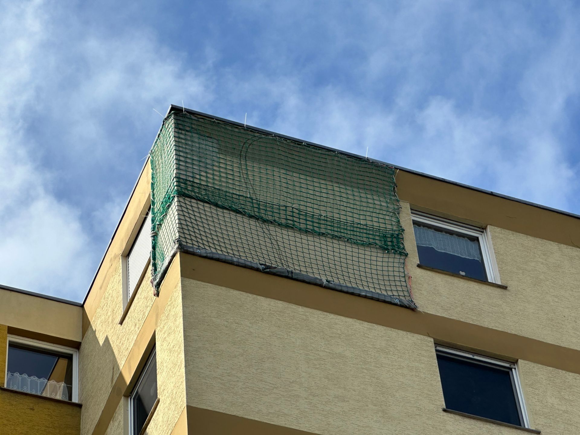Gebäudeecke mit grünem Netz, das einen Teil des Daches bedeckt. Blauer Himmel im Hintergrund.