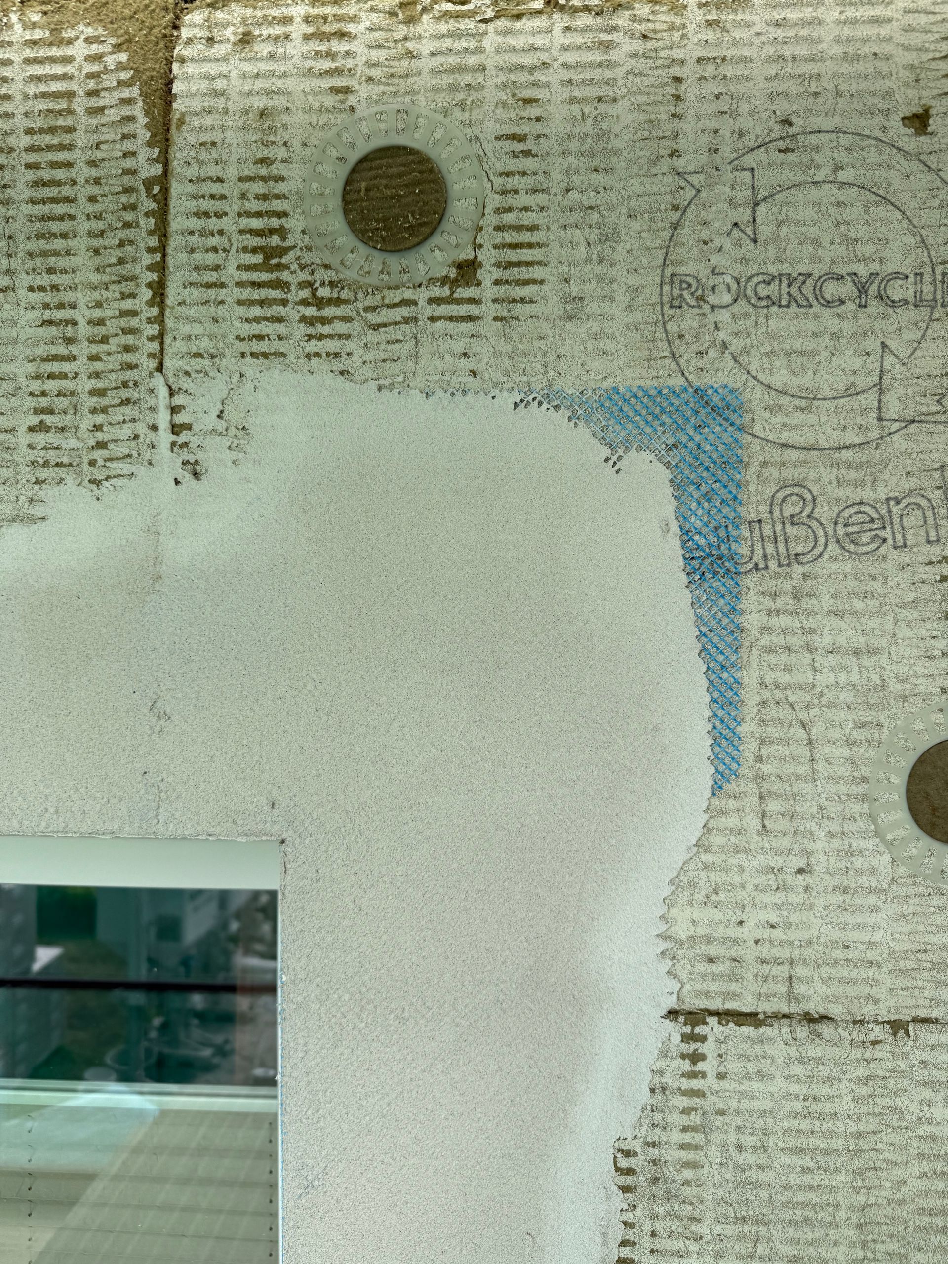 Nahaufnahme der Außenwand mit weißem Granulat, blauer Eckmarkierung und teilweise sichtbarem Fenster.