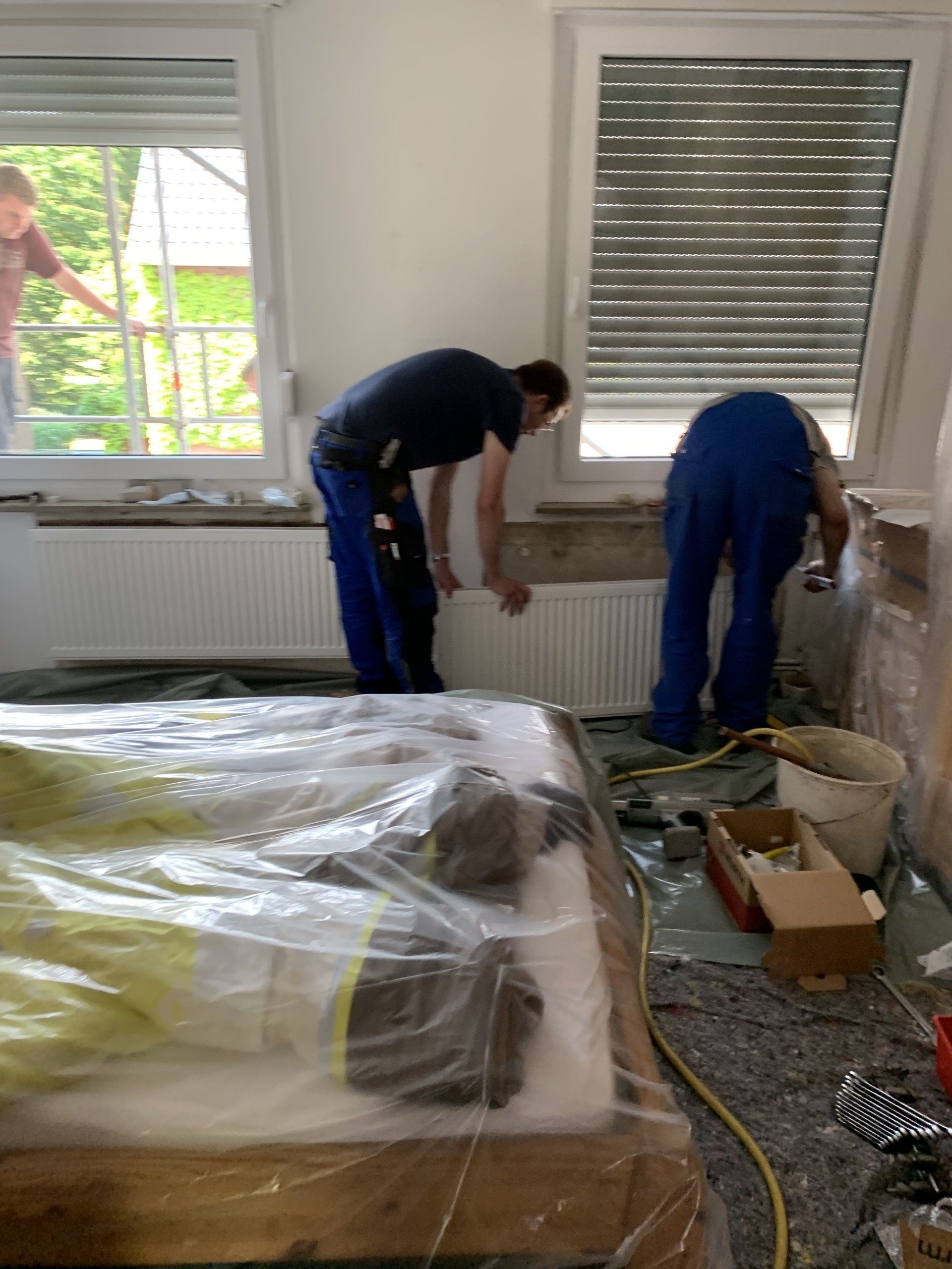 Zwei Arbeiter in blauen Overalls installieren einen Heizkörper in einem Zimmer. Ein Bett ist mit Plastikfolie abgedeckt.