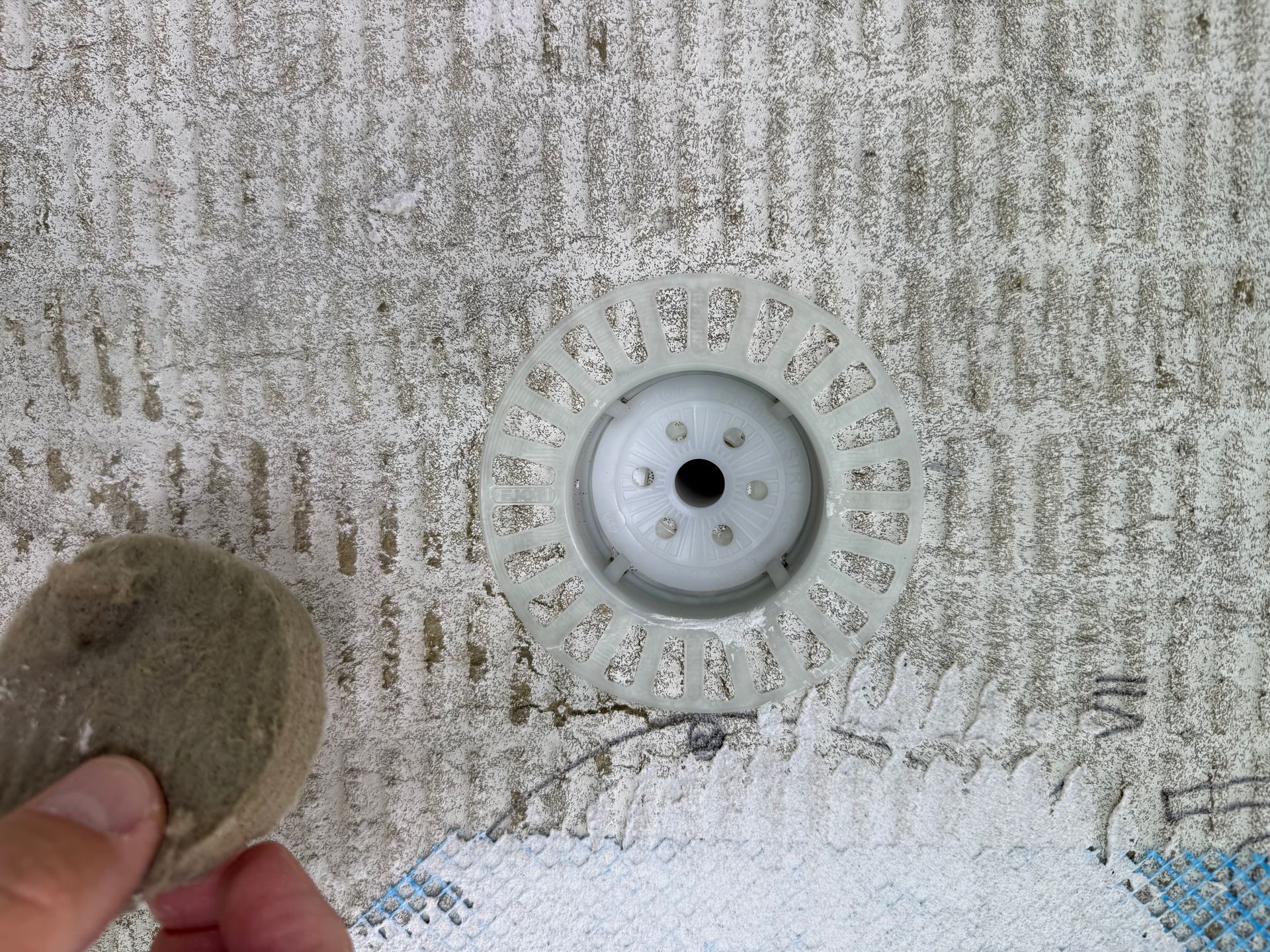 Eine Hand hält einen Stein neben einer weißen Kunststoff-Poolarmatur, die in eine strukturierte, graue Wand eingebettet ist.