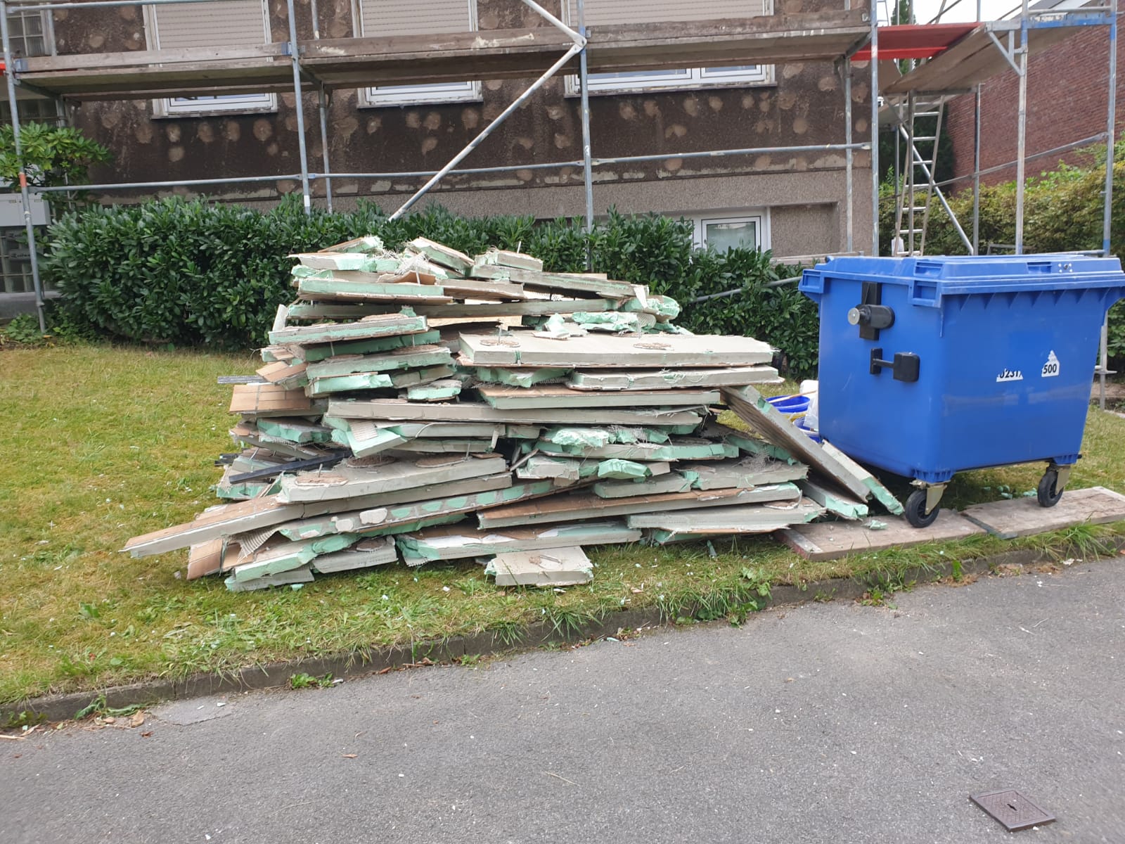 Stapel grauer Betonstücke mit grüner Isolierung neben einer blauen Mülltonne auf einer Wiese, Baustelle.