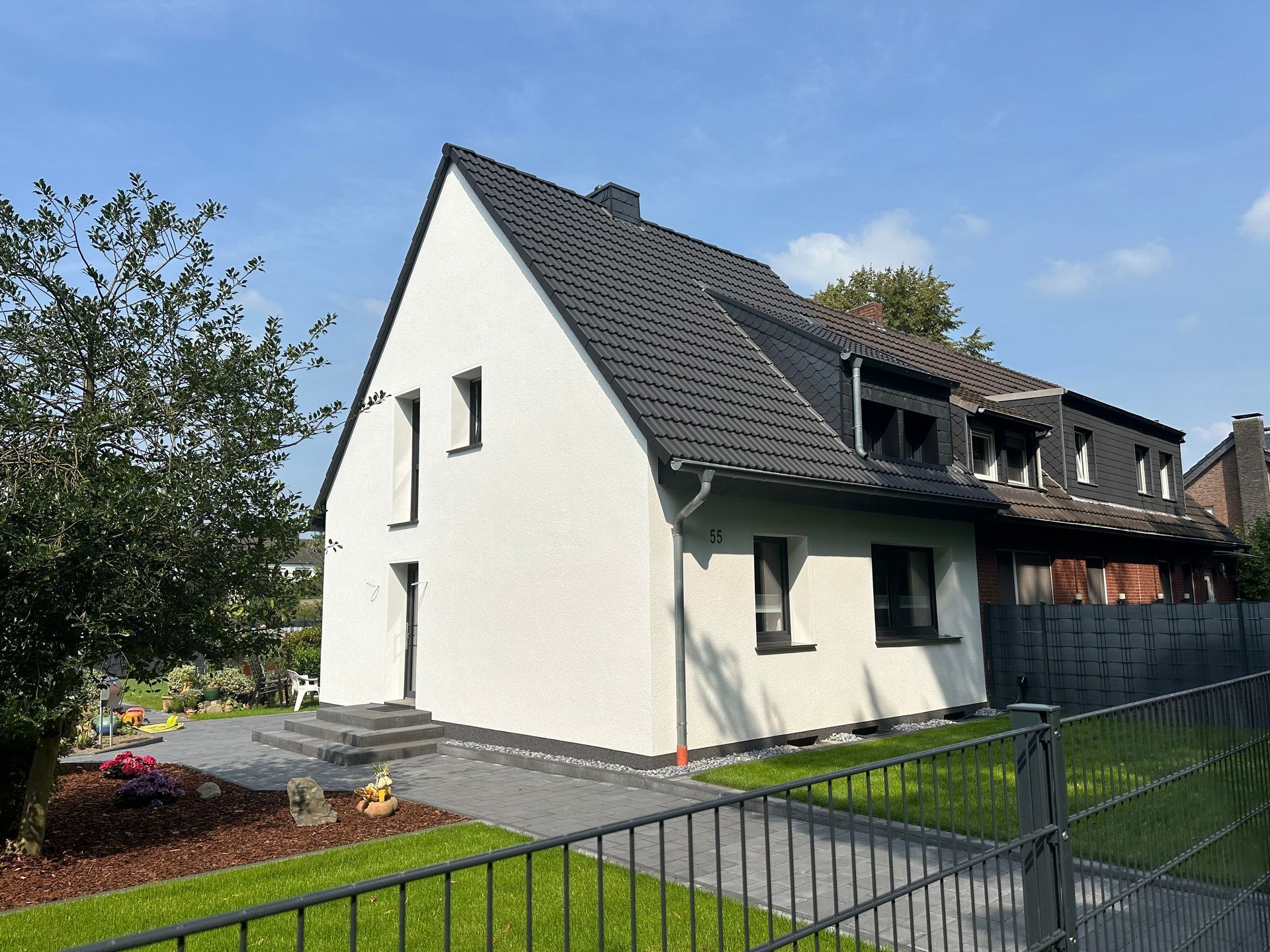 Weißes Haus mit schwarzem Dach und schwarzen Fenstern, Steinweg, eingezäunter Rasen, sonniger Tag.