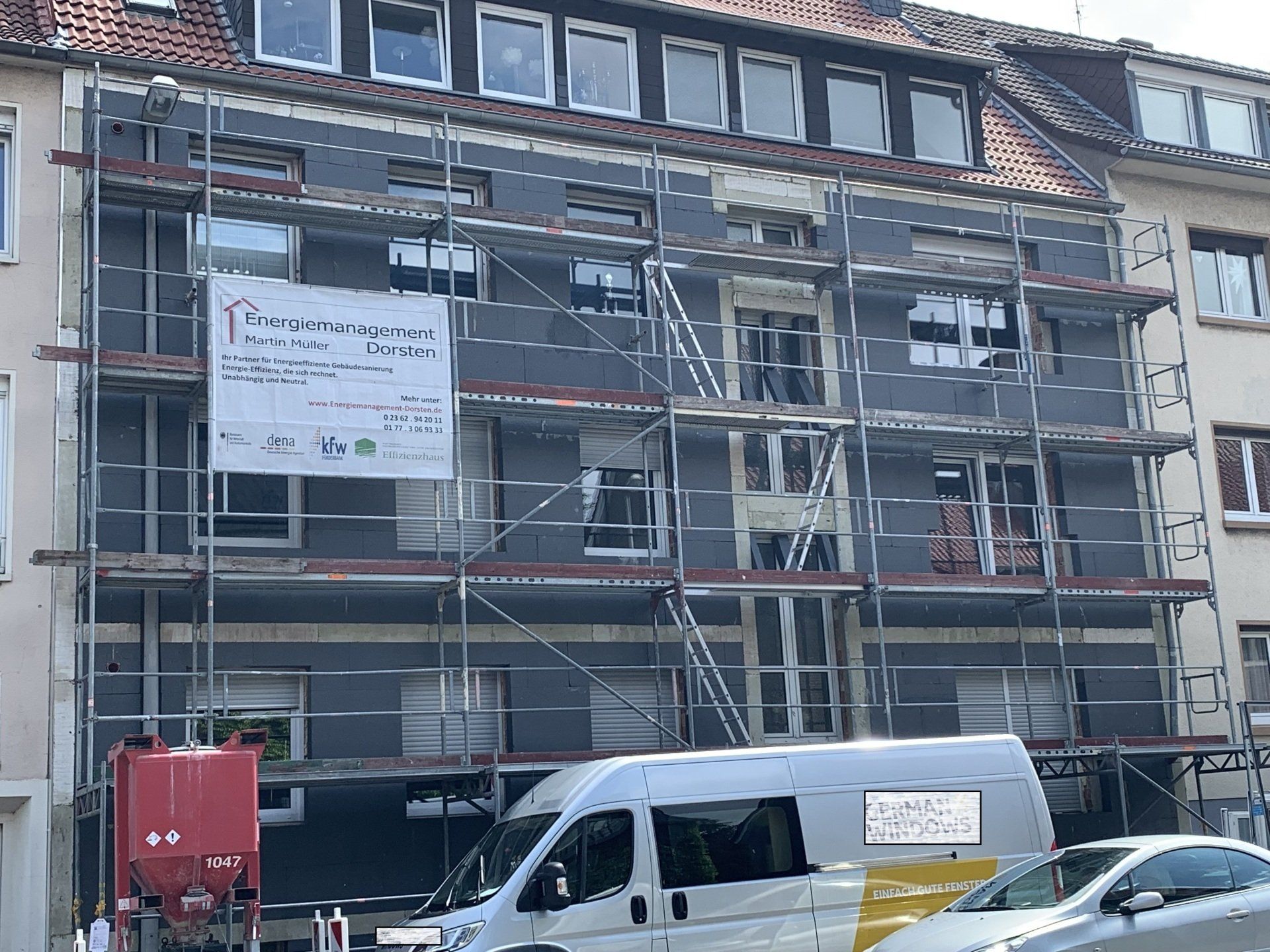 Gebäudefassade wird renoviert, mit Gerüsten und dunkelgrauer Farbe bedeckt. An der Fassade ist ein Banner sichtbar.