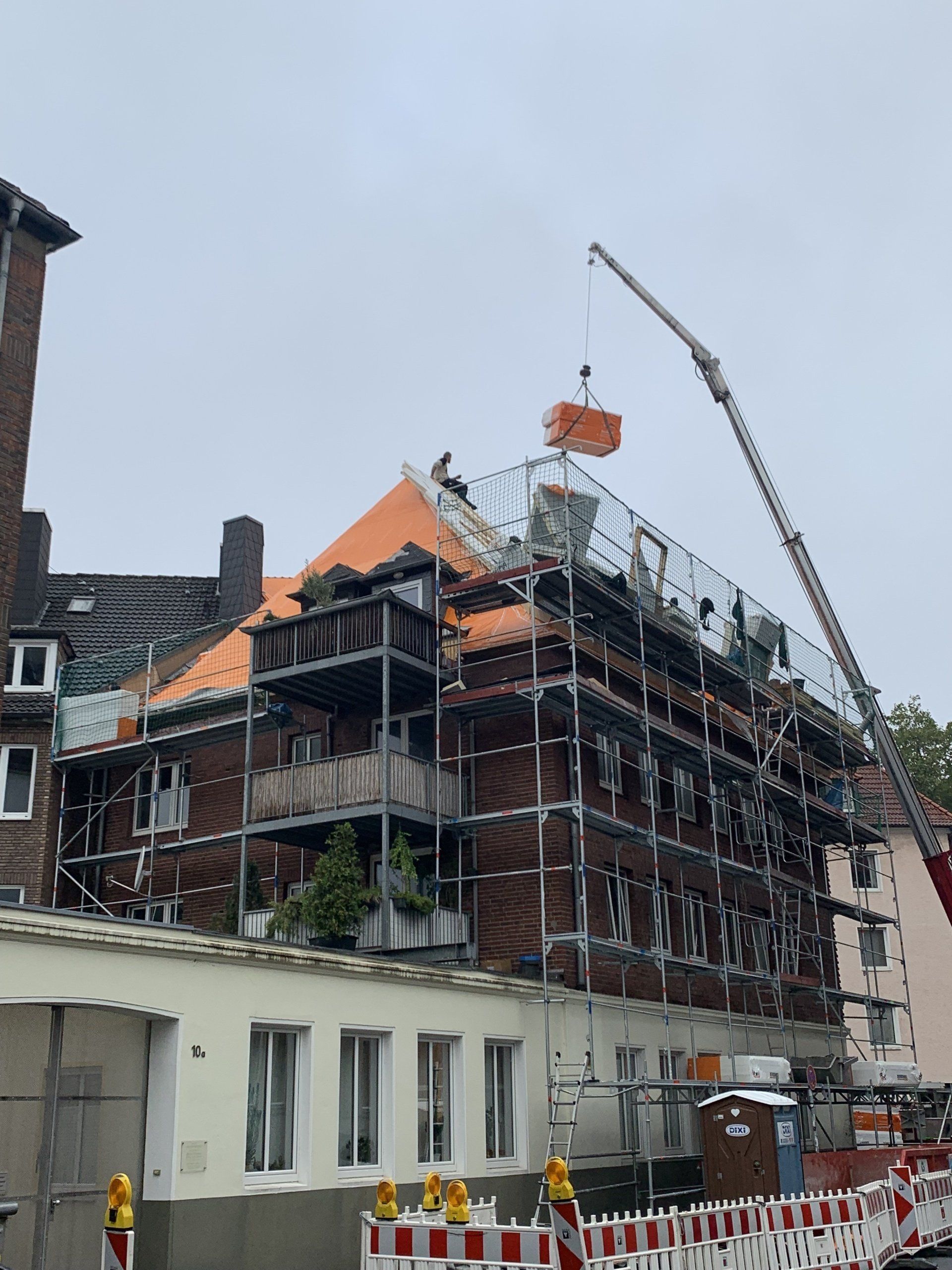 Gebäudesanierung: Ein Kran hebt einen orangefarbenen Container vom Dach, ein Gerüst umgibt ein Backsteingebäude.