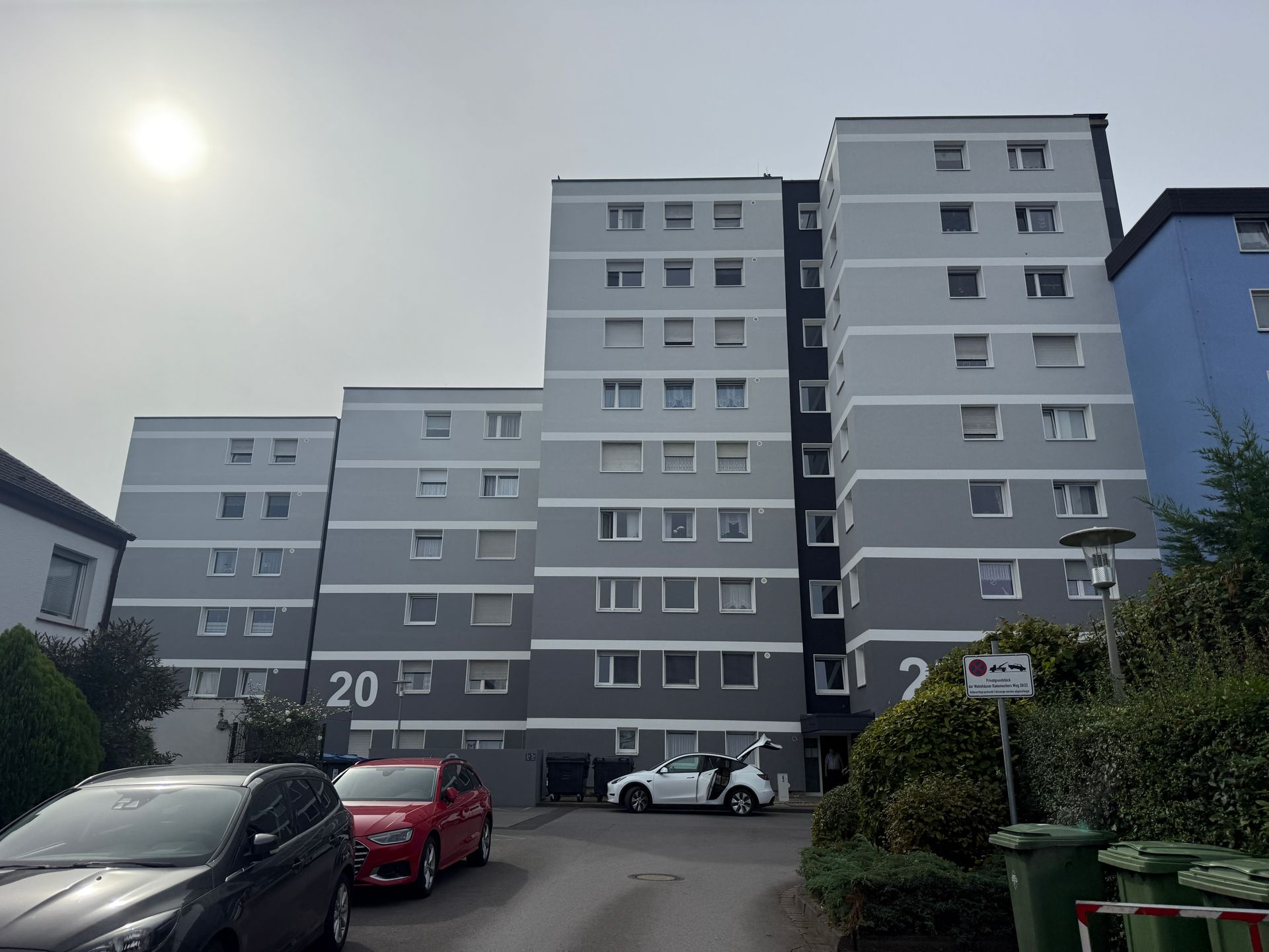 Wohnhäuser mit grauer Fassade; davor parkende Autos, bewölkter Himmel darüber.