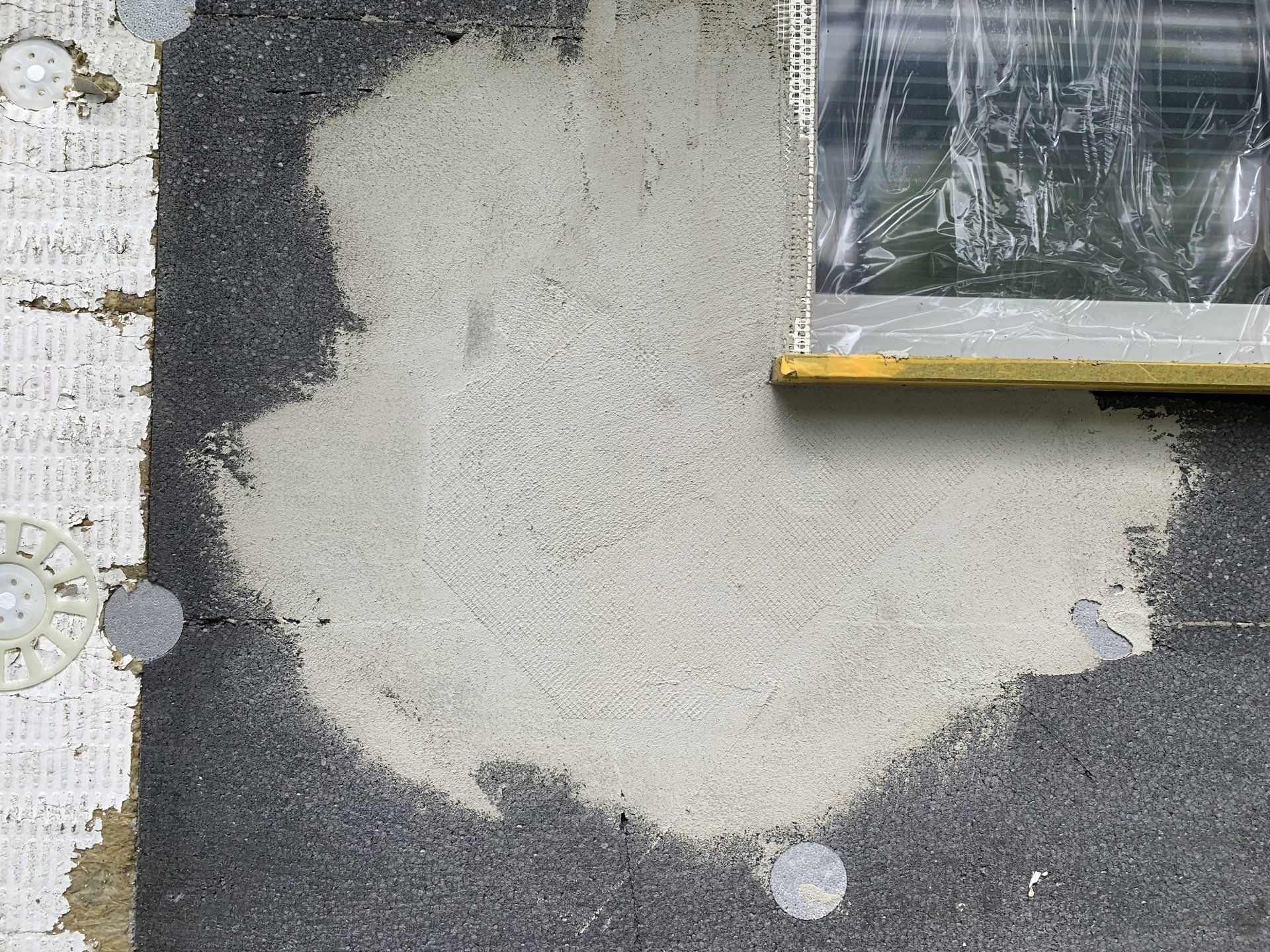 Ein grauer Mörtelfleck auf dunkler Isolierung, in der Nähe eines mit Plastik abgedeckten Fensters.