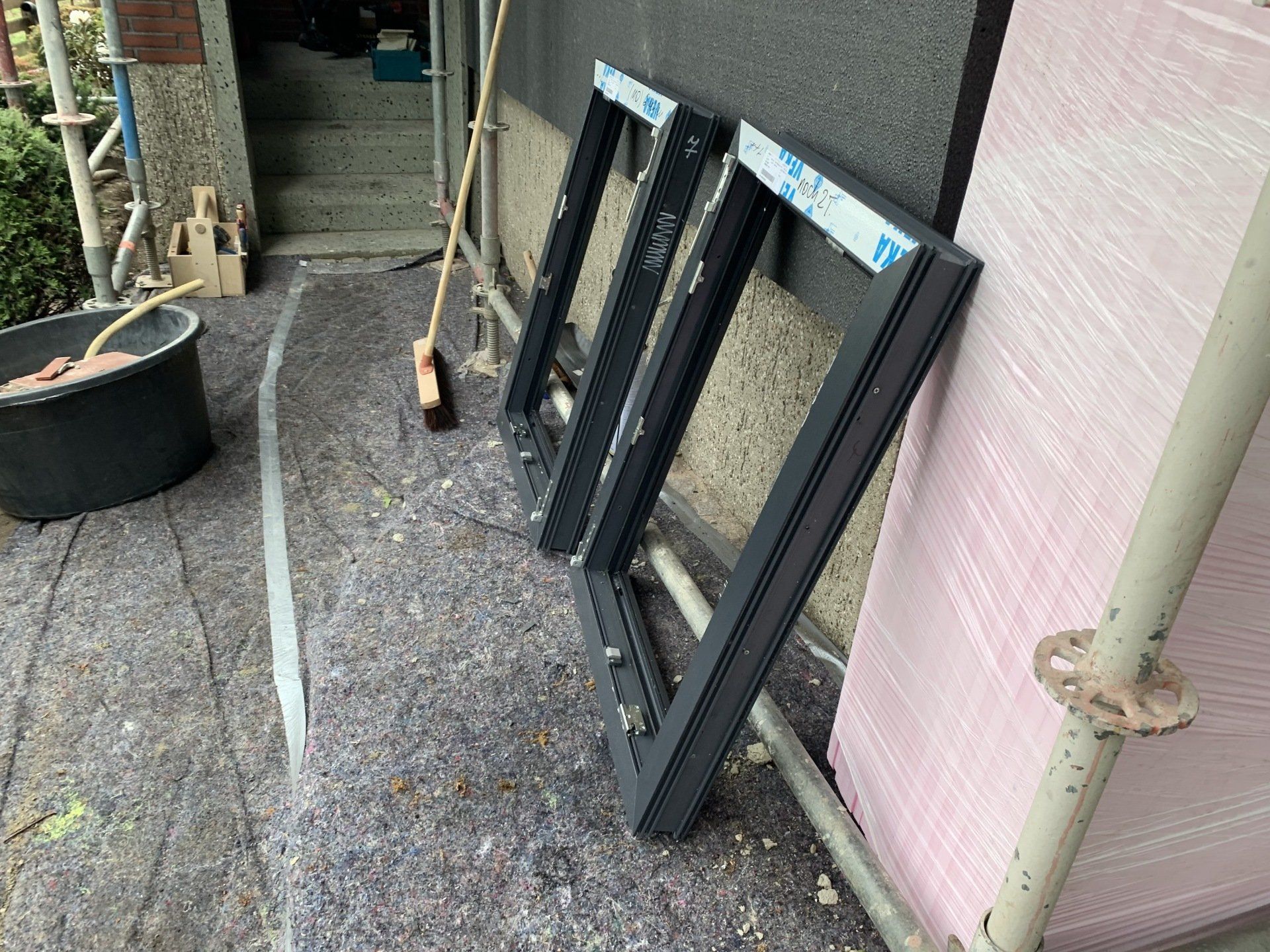 Zwei dunkle Fensterrahmen, die an eine rosa Isolierung gelehnt sind, in der Nähe eines Gerüsts.