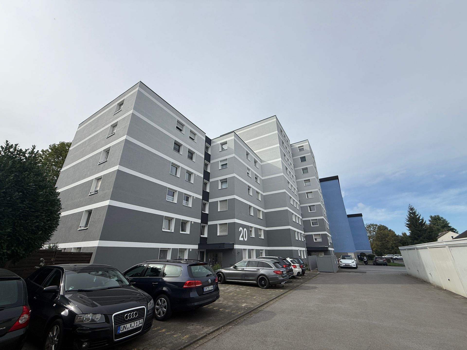 Mehrstöckiges grau-weißes Wohnhaus mit davor geparkten Autos unter einem bewölkten Himmel.