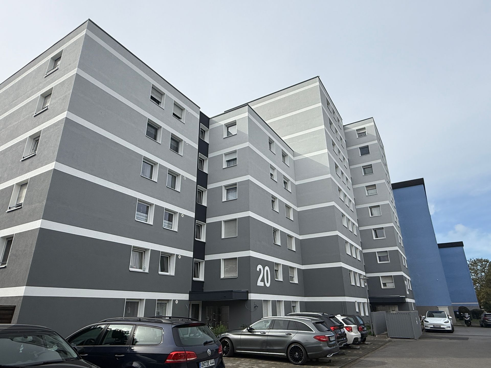 Grau-weißes Wohnhaus mit davor geparkten Autos. Über dem Eingang ist die Nummer „20“ zu sehen.