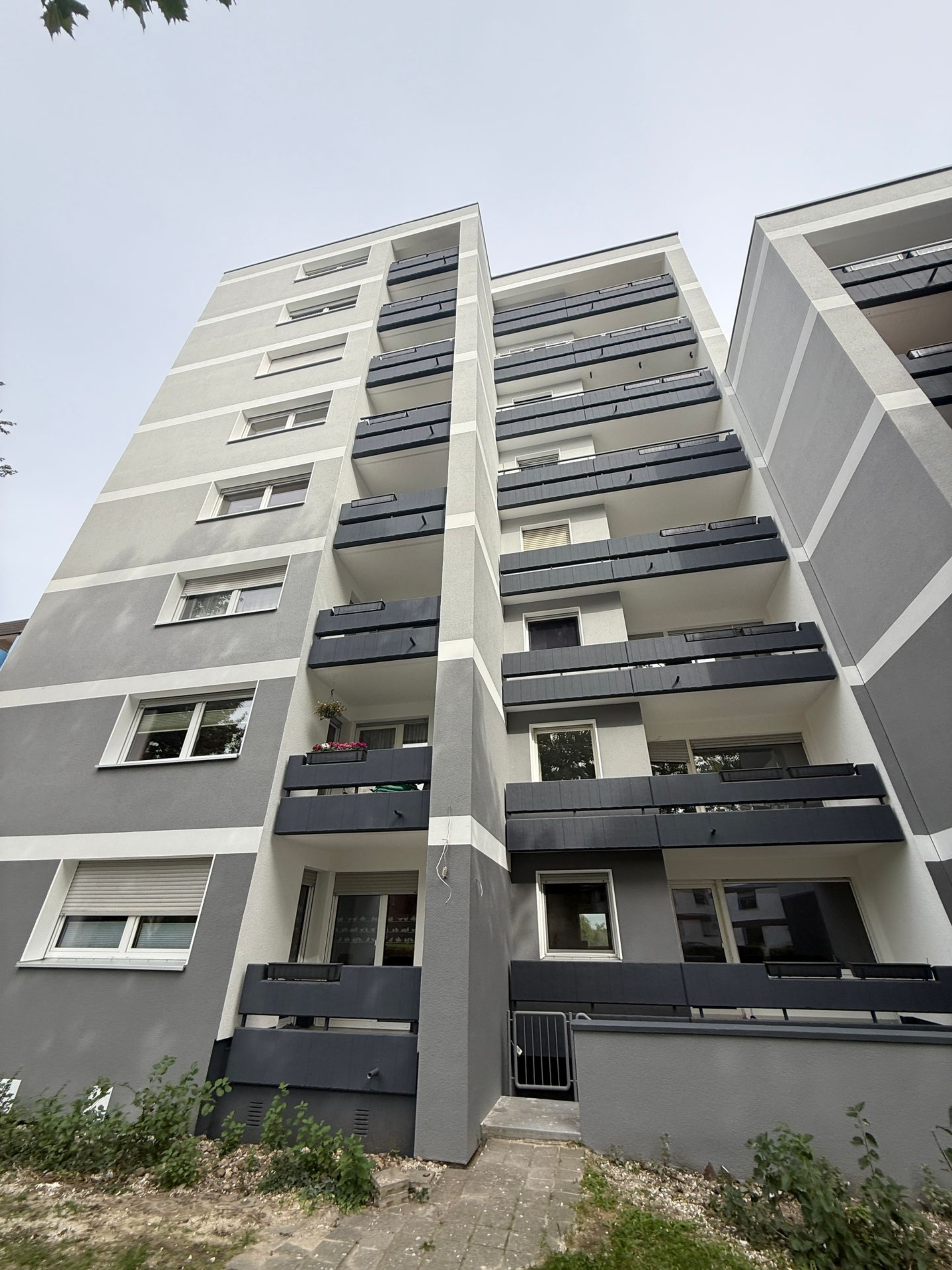 Modernes mehrstöckiges Gebäude mit grau-weißer Fassade, schwarzen Fensterabdeckungen und vielen Fenstern.