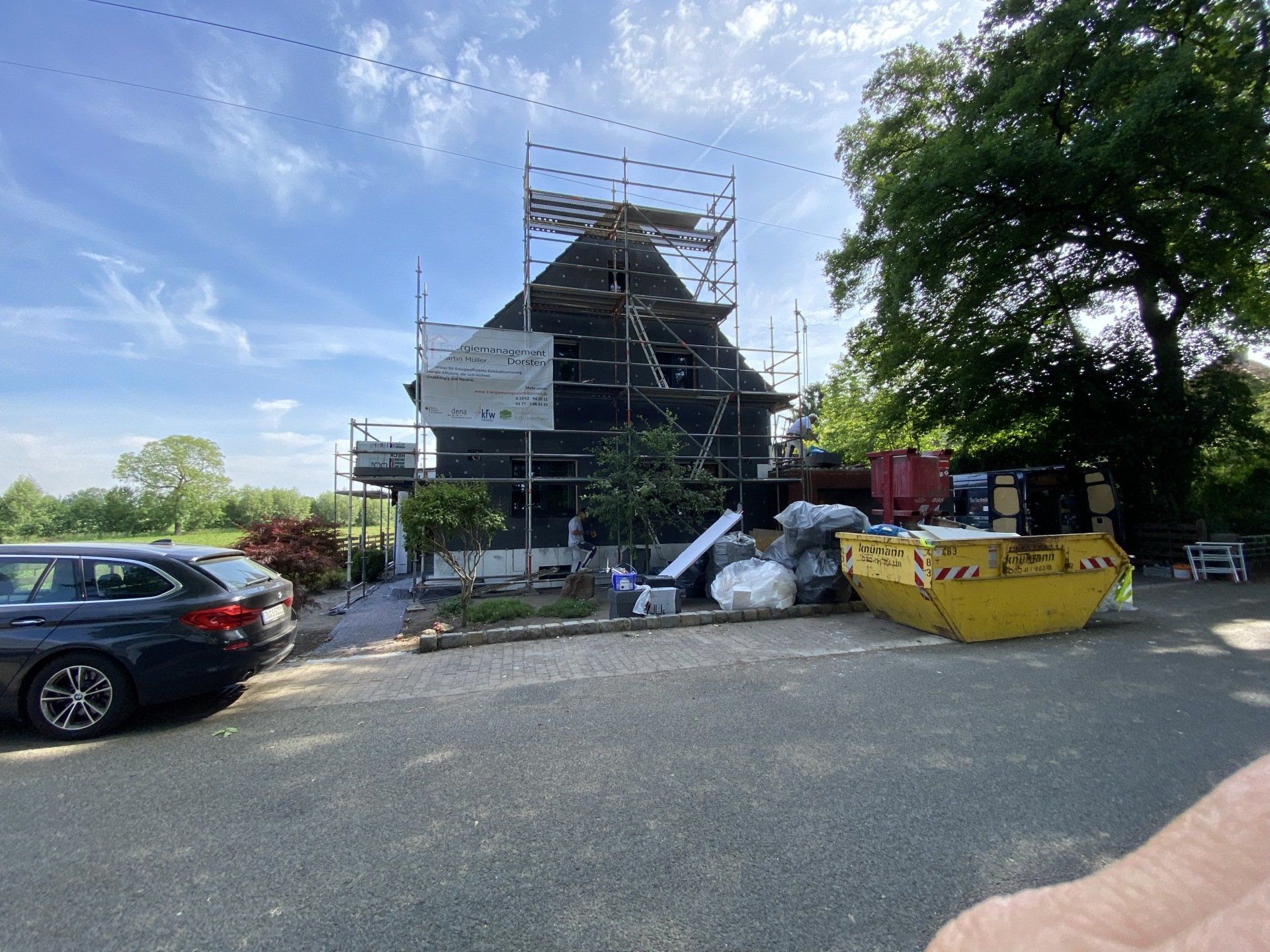 Haus im Bau mit Gerüst, blauer Himmel, gelber Müllcontainer, geparktes Auto.