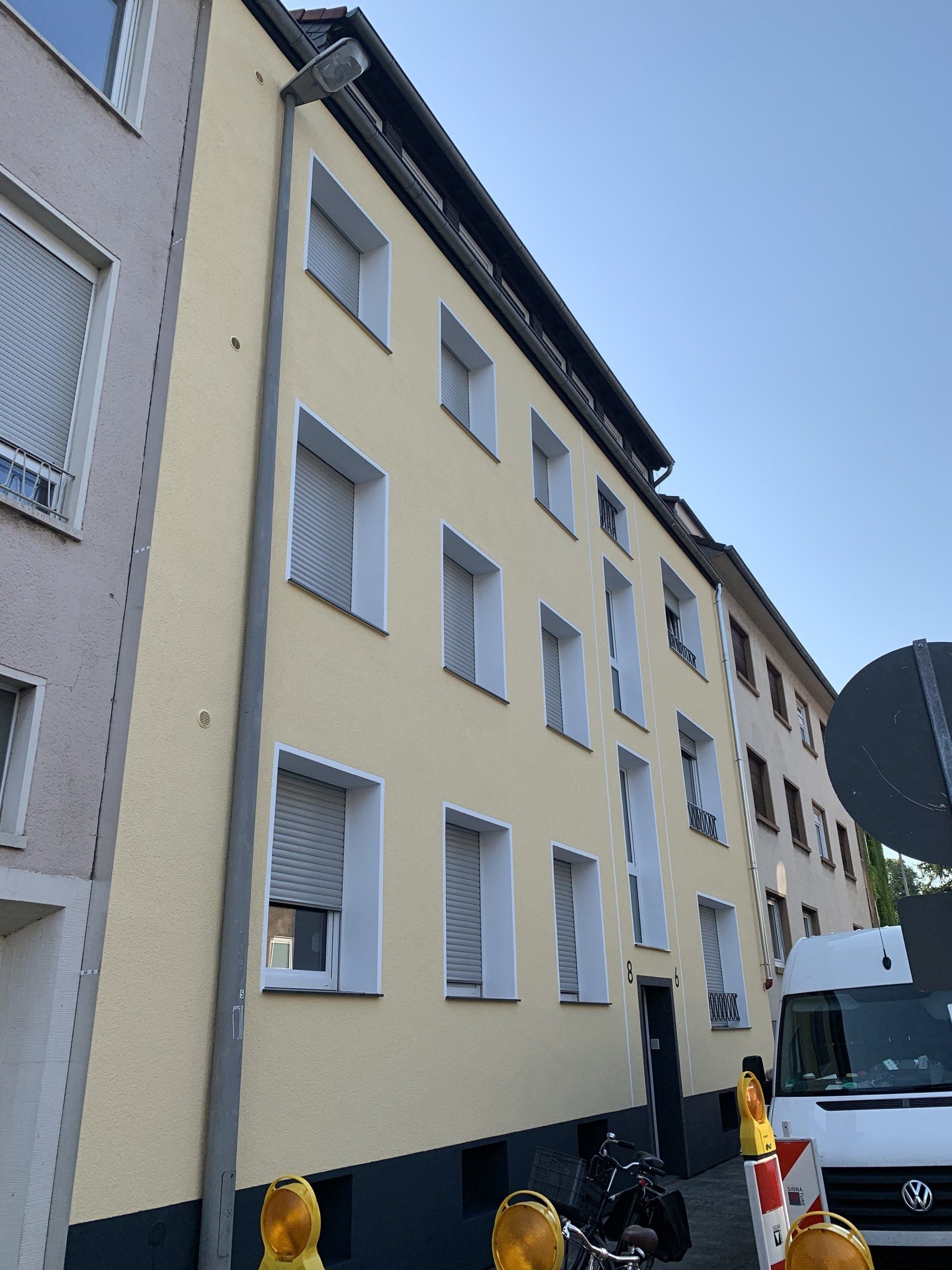 Beiges Wohnhaus mit mehreren Fenstern, Dachrinne an der Seite und Bauabsperrungen davor.