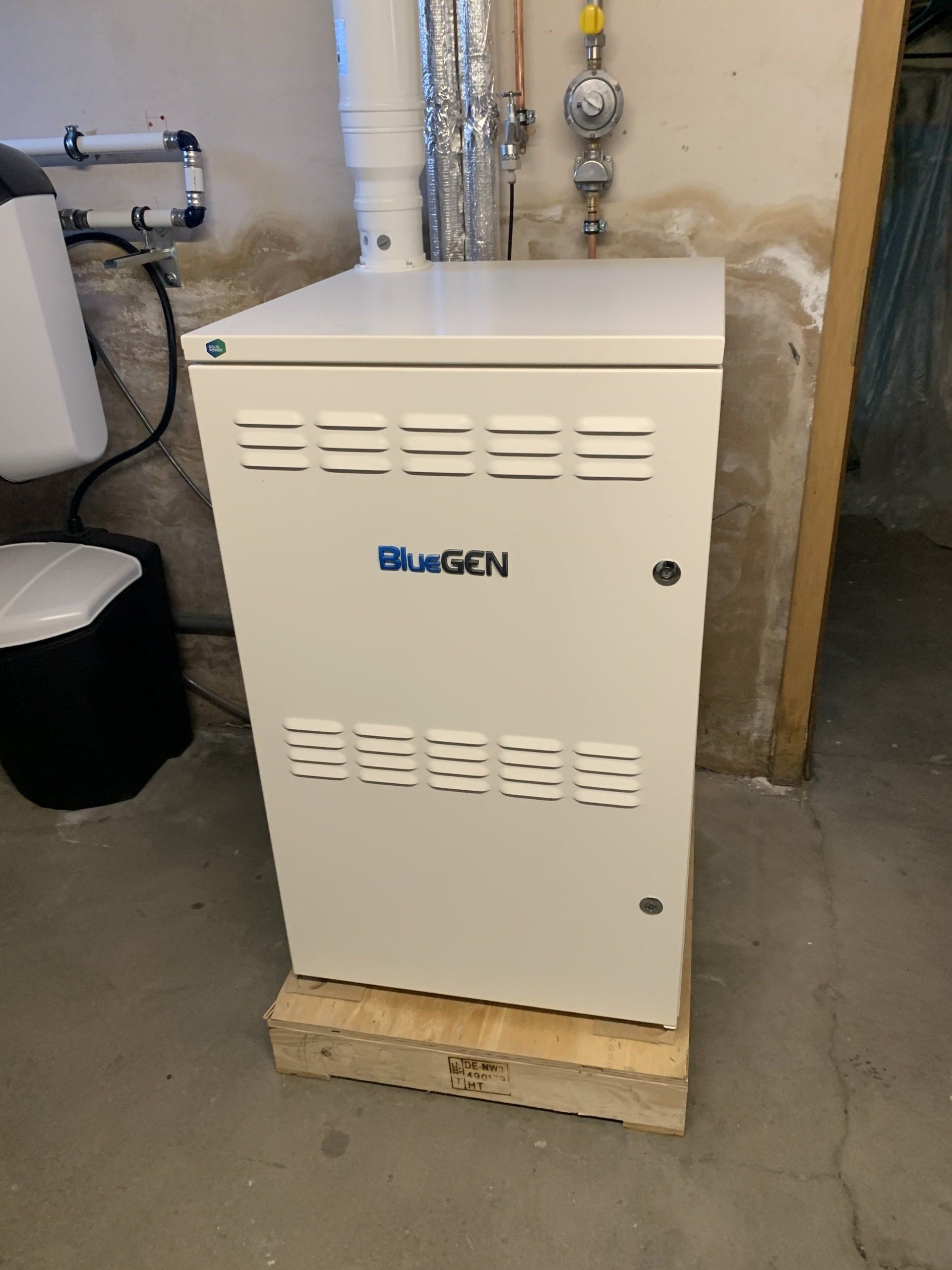 Weißer BlueGen-Generator auf einer Holzplattform in einem Keller. Dahinter sind Gasleitungen sichtbar.