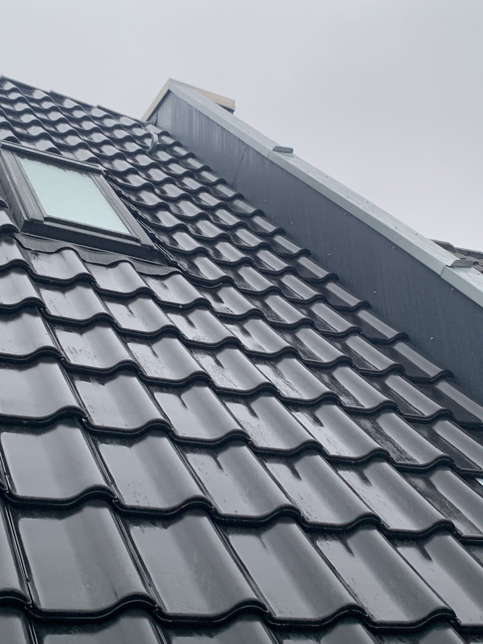 Schwarzes Ziegeldach mit Oberlicht und Regenrinne unter bewölktem Himmel.