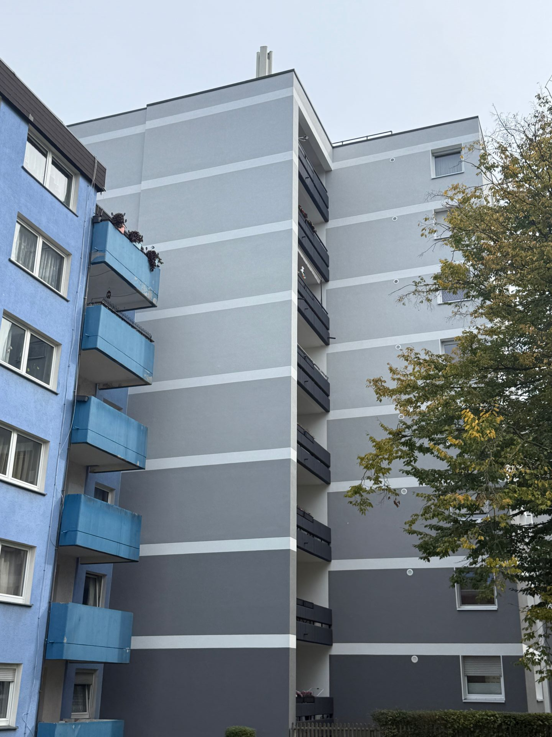 Mehrstöckiges Gebäude mit unterschiedlichen Grau- und Blautönen an der Außenseite. Grüne Bäume im Vordergrund.