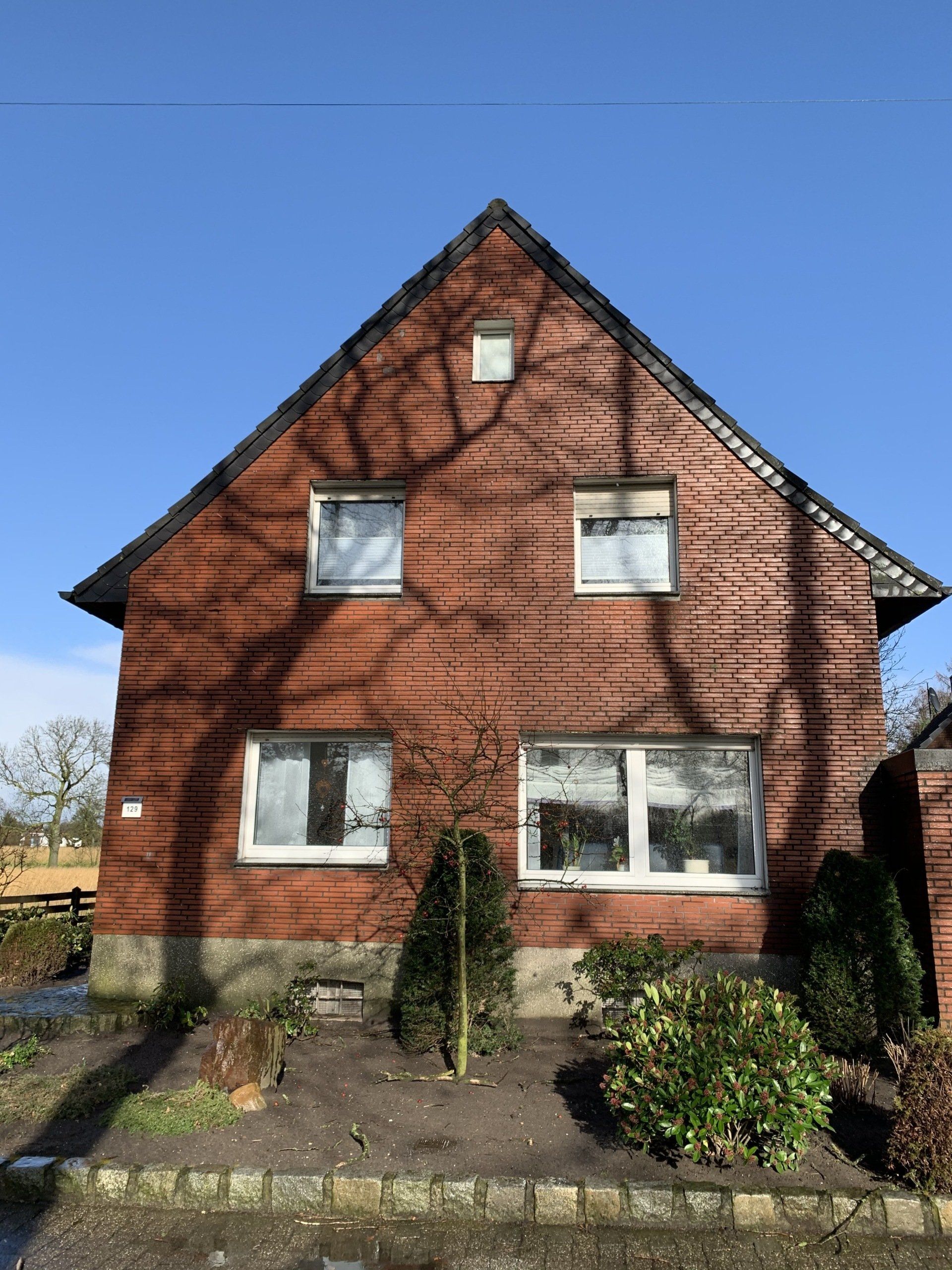 Zweistöckiges Backsteinhaus mit Satteldach, eingerahmt von Baumschatten vor blauem Himmel.