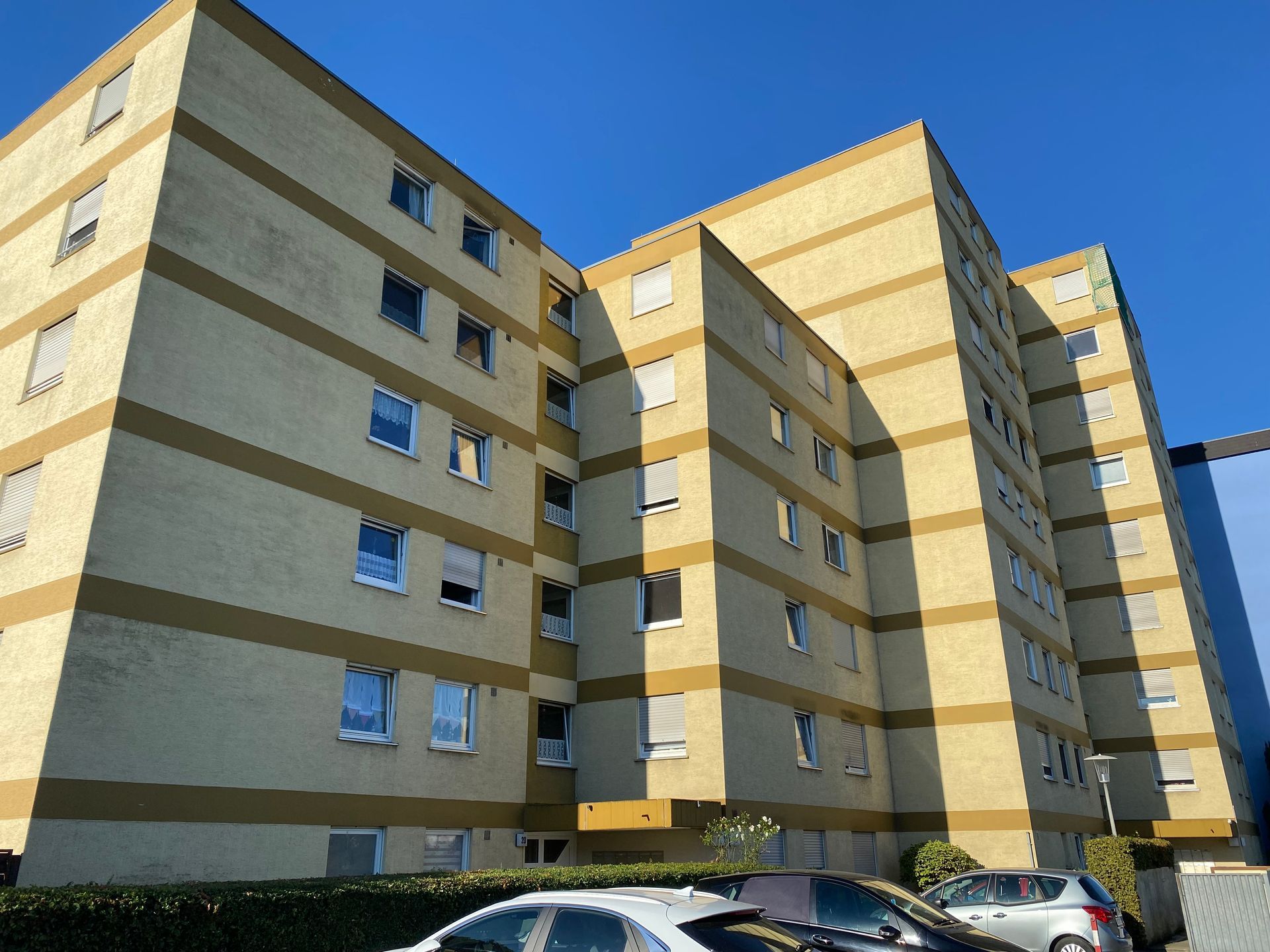 Gelbes Wohnhaus mit dunkelbraunen Querstreifen, blauer Himmel, davor parkende Autos.