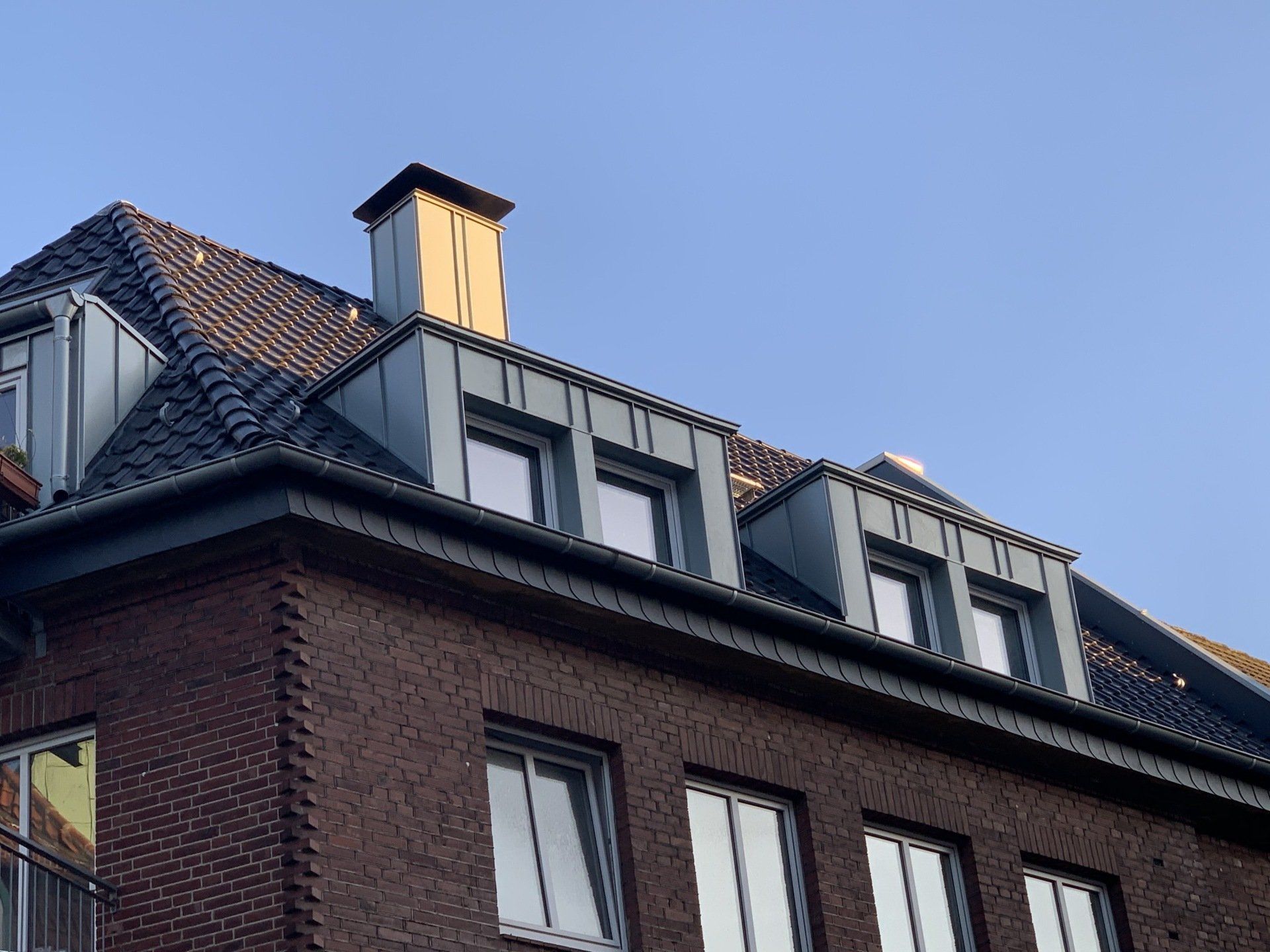 Backsteingebäude mit dunklem Ziegeldach, Dachgauben, Fenstern und einem Schornstein vor blauem Himmel.