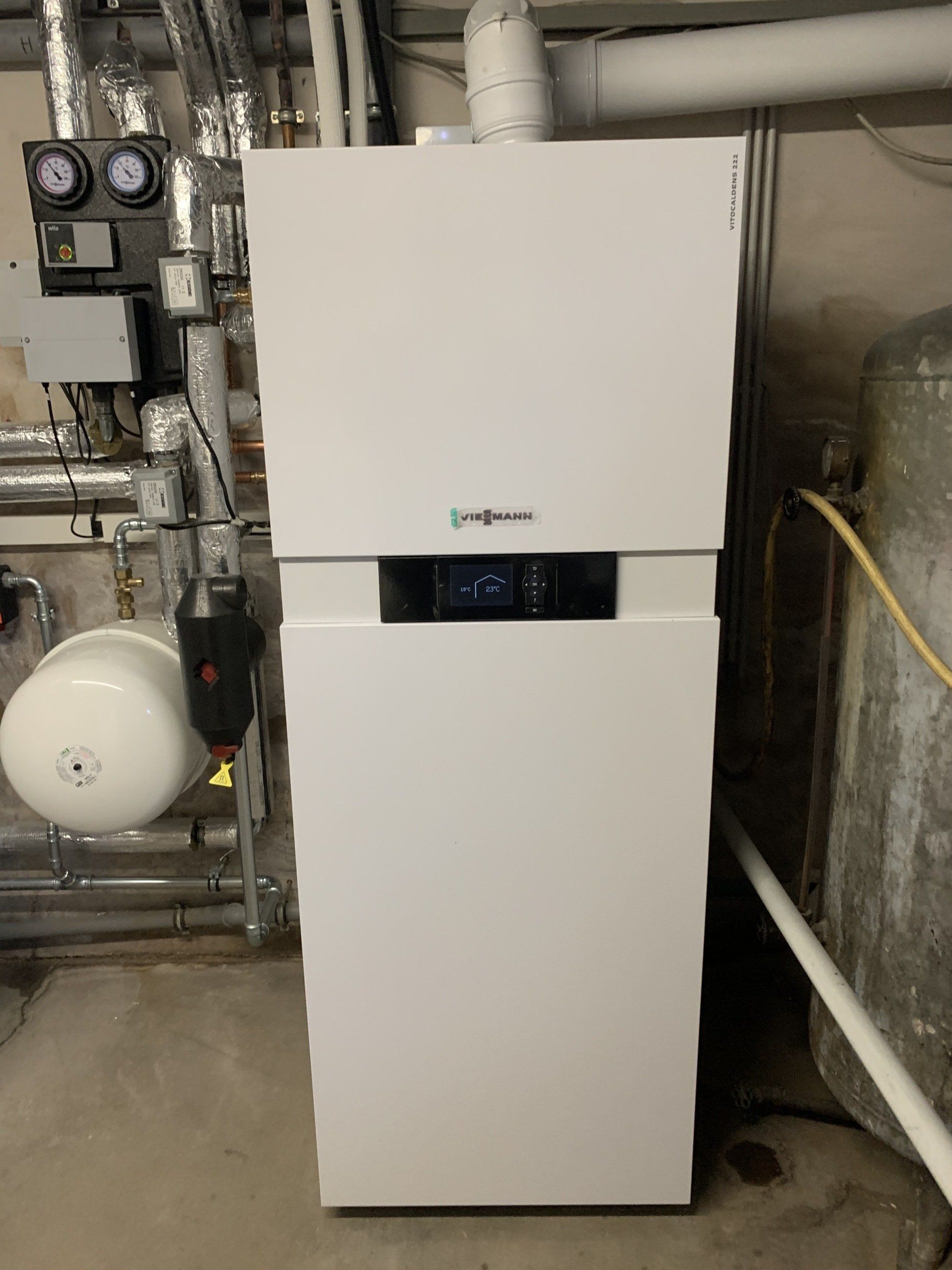 Weißes Heizsystem in einem Keller mit Rohren, Messgeräten und einem Wassertank.