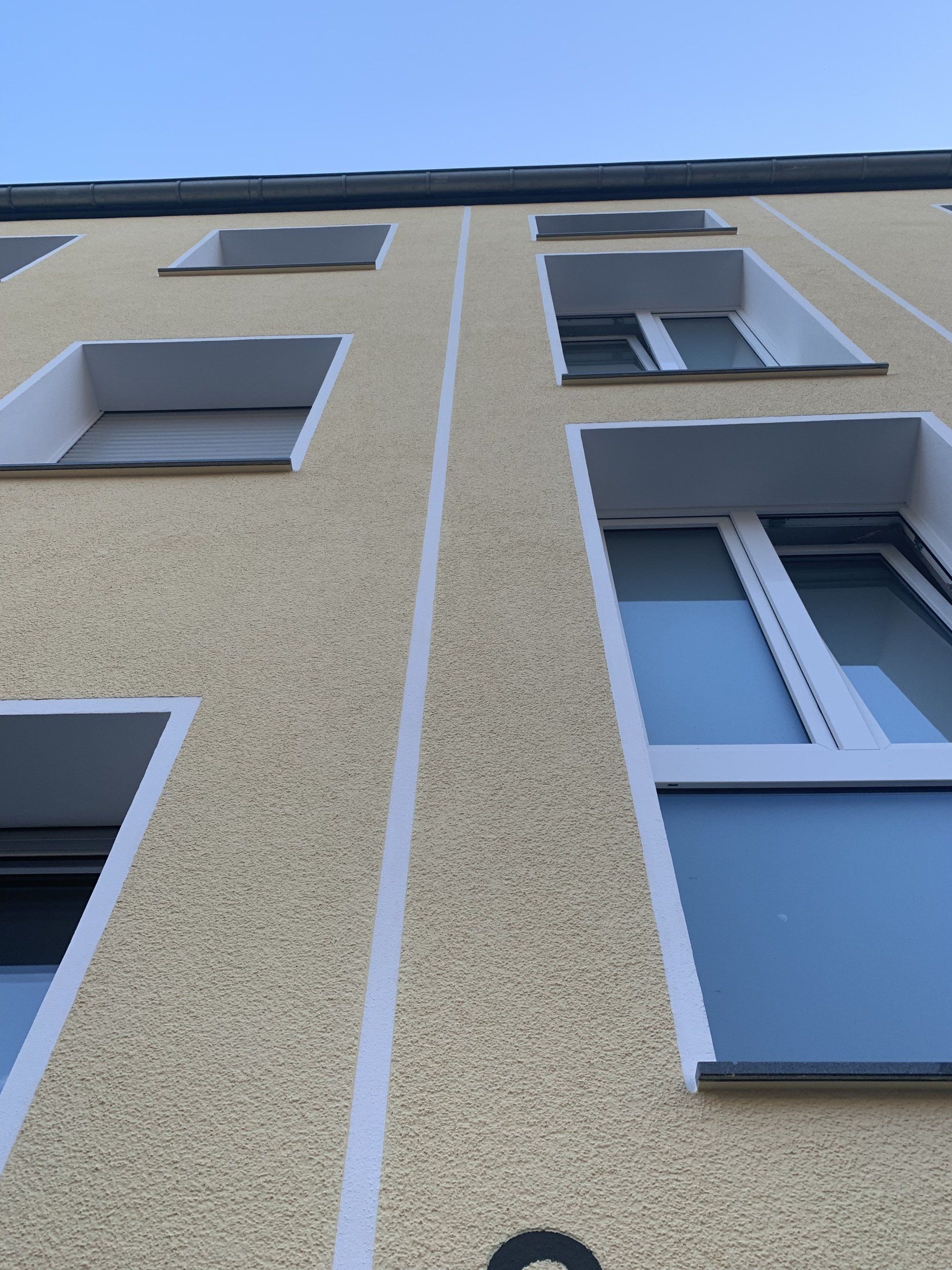 Gelbes Gebäude mit weiß gerahmten Fenstern und vertikalen weißen Streifen vor blauem Himmel.