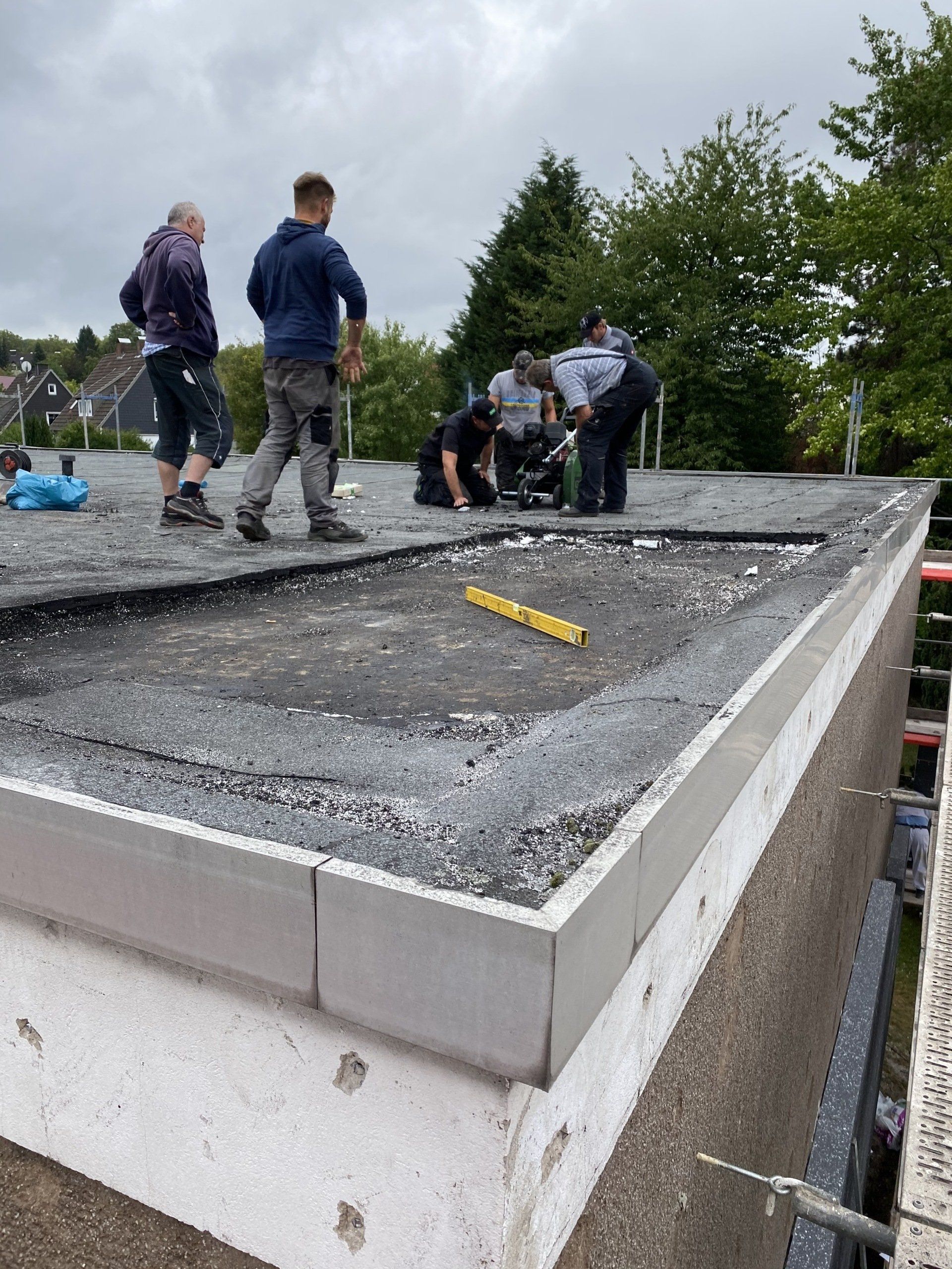 Bauarbeiter auf einem Flachdach installieren eine Dacheinfassung. Graue, schwarze und weiße Materialien. Bewölkter Tag.