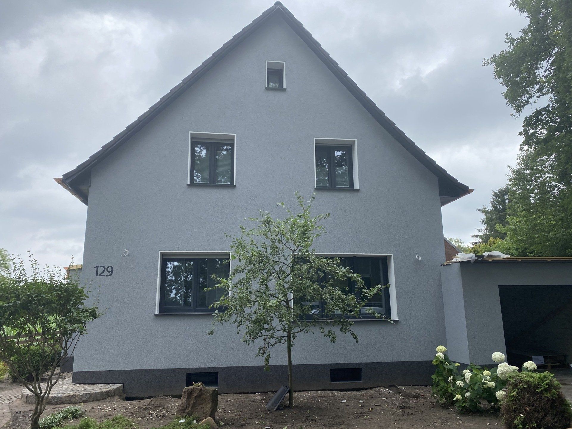 Graues zweistöckiges Haus mit schwarz gerahmten Fenstern und einem kleinen Baum davor, Adresse 129.