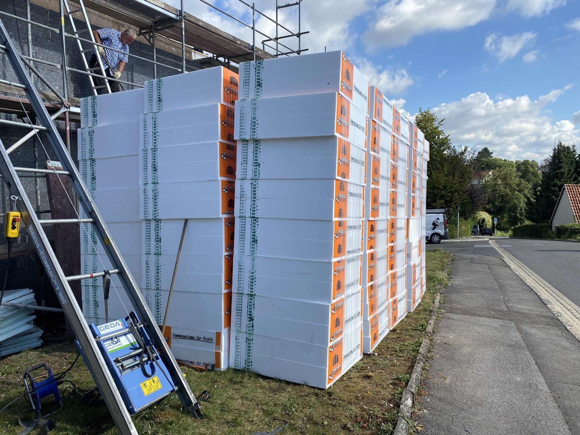 Stapel weißer Dämmplatten auf einer Wiese neben einem Gebäude, mit einem Arbeiter auf einem Gerüst; sonniger Tag.