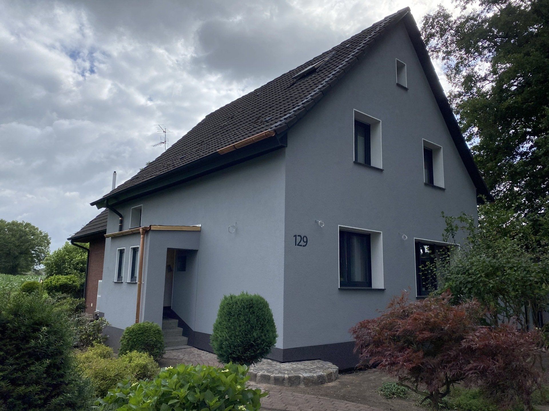 Graues Haus mit dunklem Dach und Zierleisten, umgeben von Grün unter einem bewölkten Himmel.