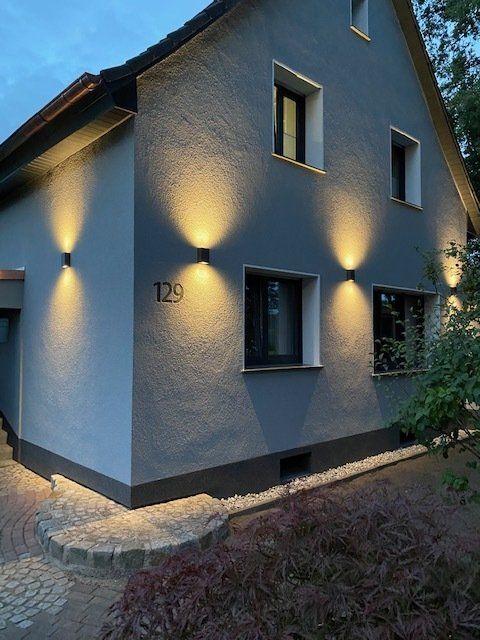 Graues Haus mit Up-and-Down-Wandleuchten, die die Fassade beleuchten. Die Hausnummer „129“ ist sichtbar.