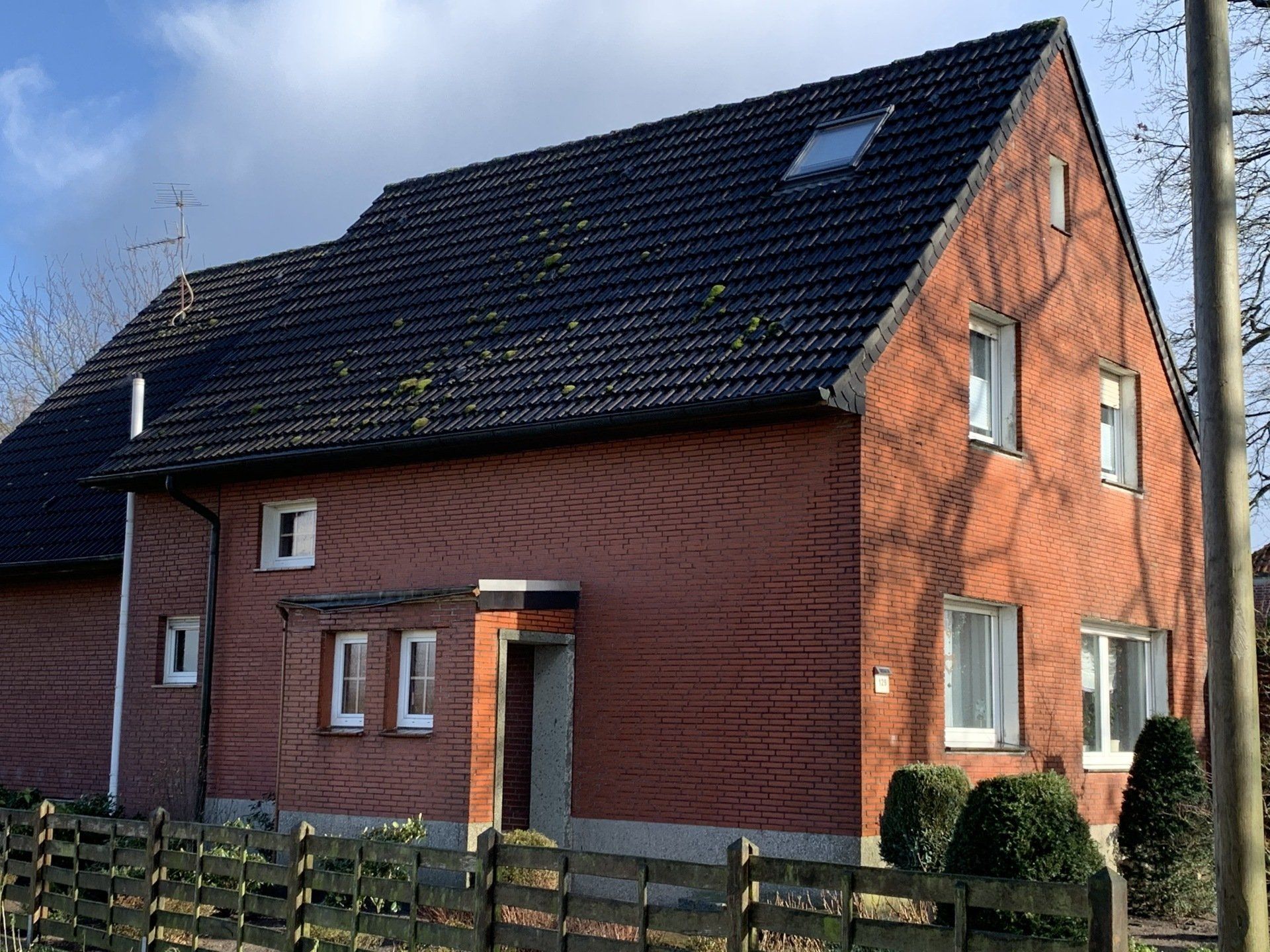 Rotes Backsteinhaus mit schwarzem Dach, Fenstern und einem kleinen Zaun. Sonniger Tag.
