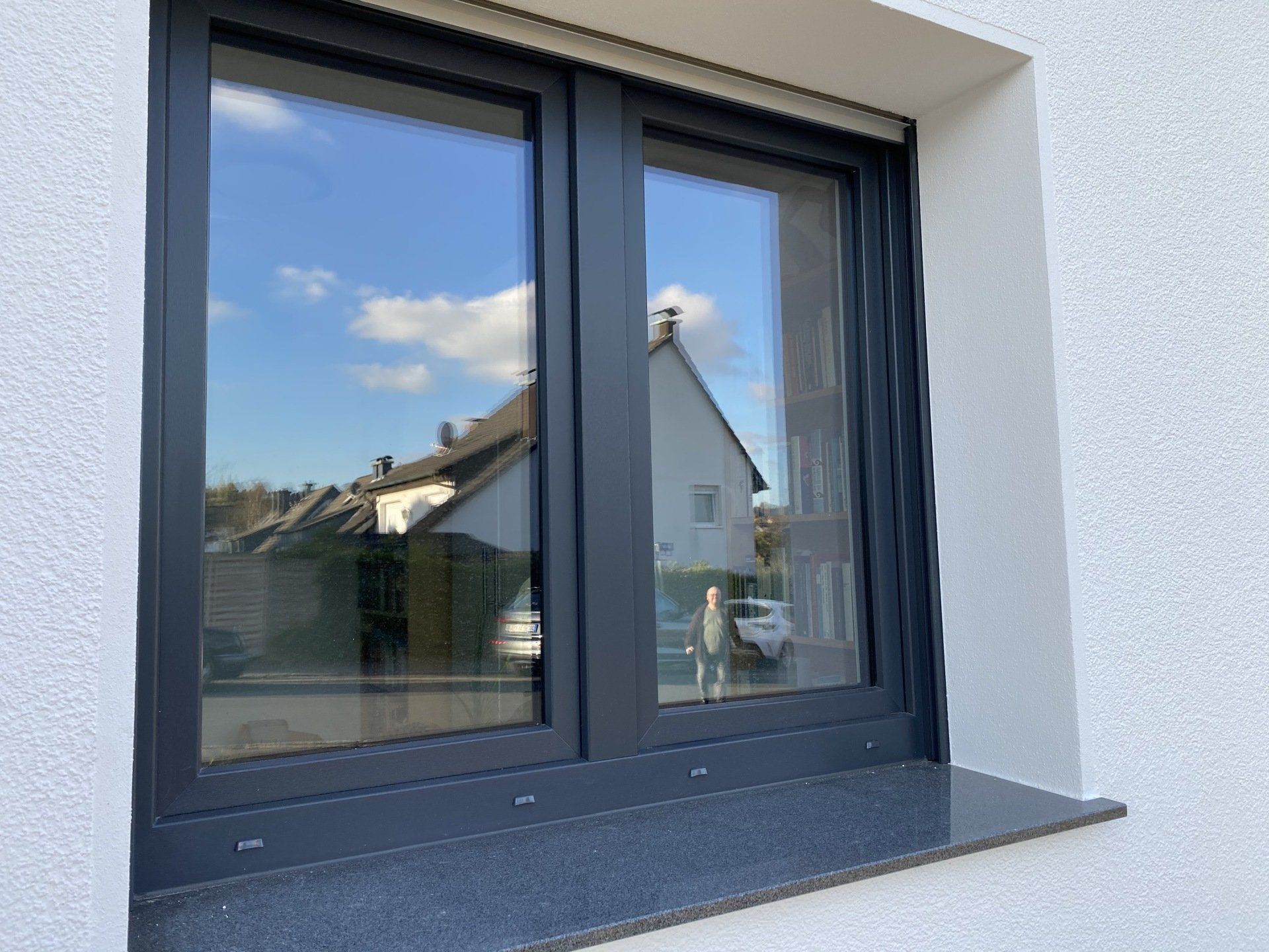 Dunkel gerahmtes Fenster, in dem sich ein Haus und ein Himmel mit Wolken spiegeln. 