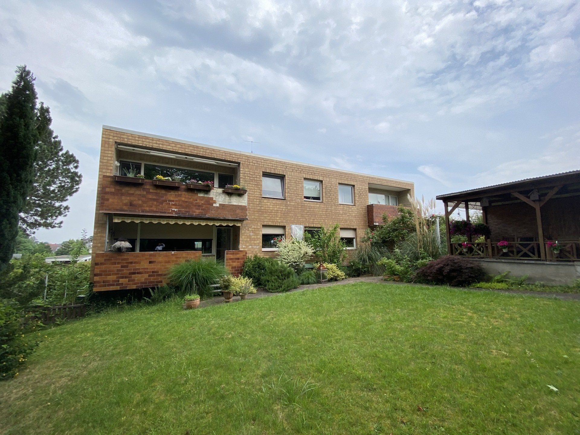Braunes und hellbraunes Backsteingebäude mit Blumenkästen, grünem Rasen, bewölktem Himmel.