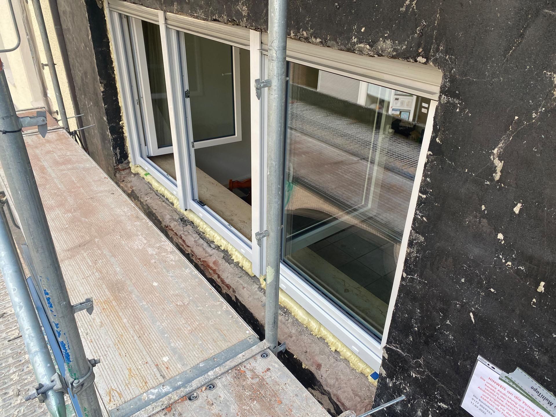 Fenster mit weißem Rahmen und geschlossenem Rollladen, von einem Außengerüst aus gesehen. Schwarze Außenwand.